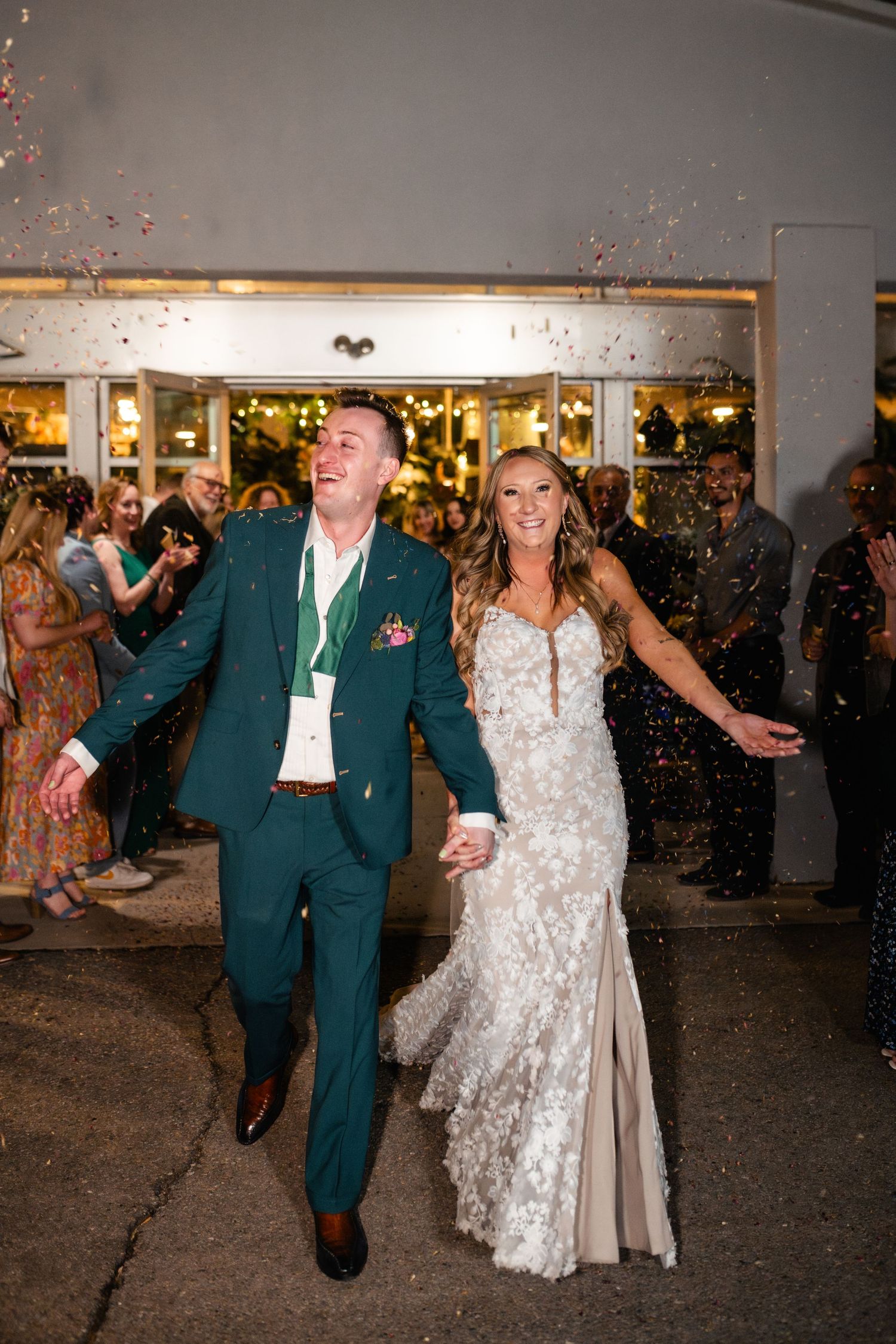 Celebrating couple exits their wedding venue amid floating confetti while wearing a green suit and white lace gown.