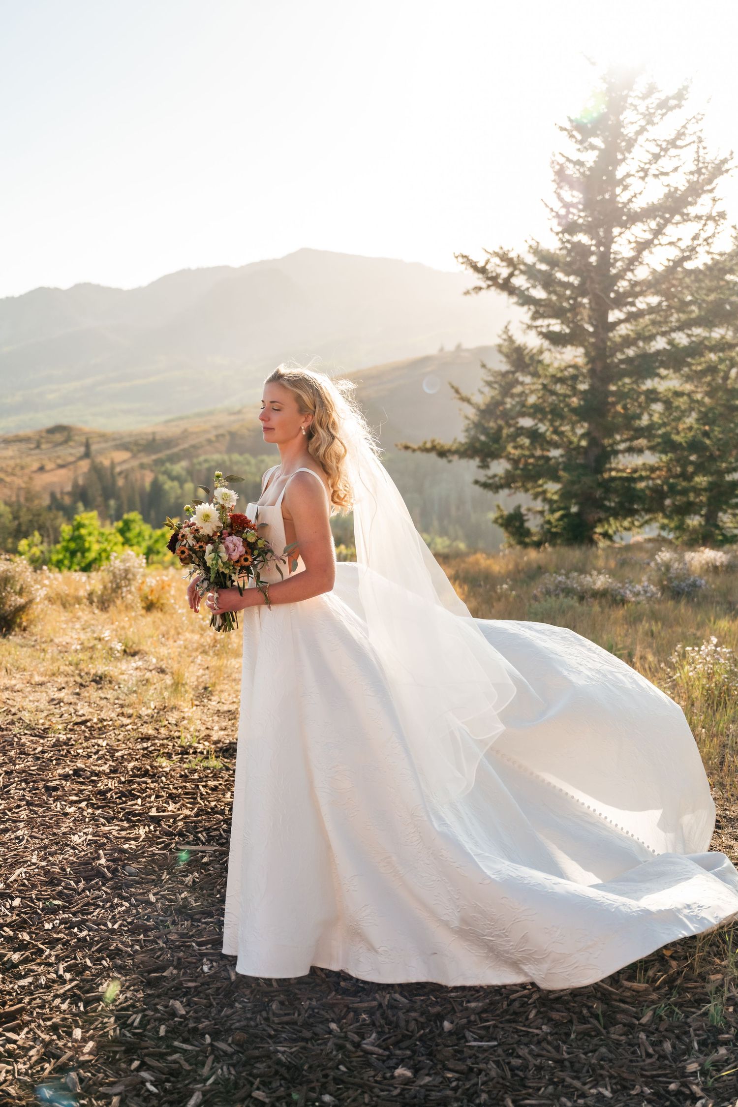 Bride in flowing white dress poses against mountain backdrop during golden hour. Park City Wedding Photographer. Utah Wedding Photographer.