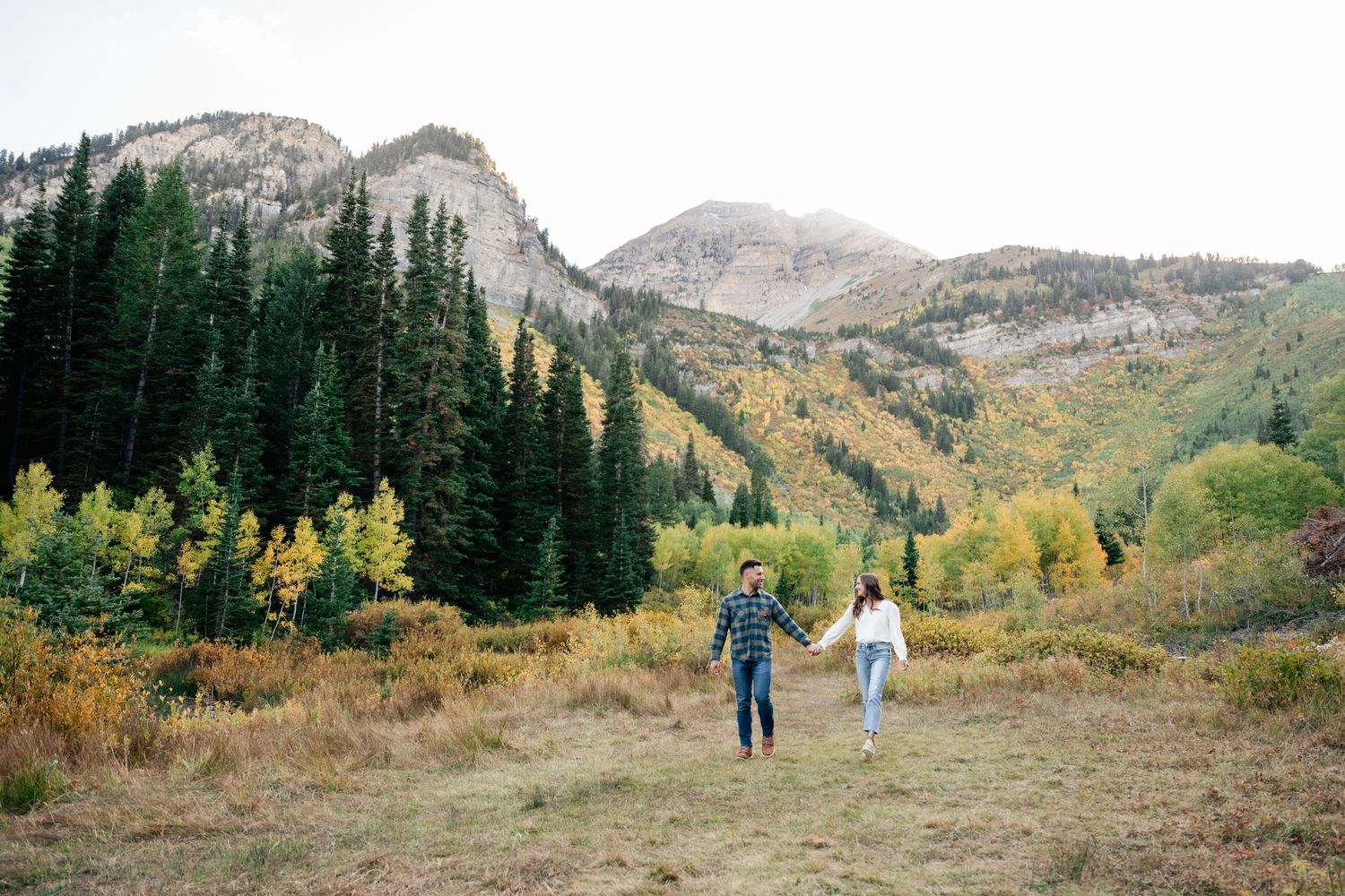 Two people walking through mountain meadow with peaks and autumn foliage in background.