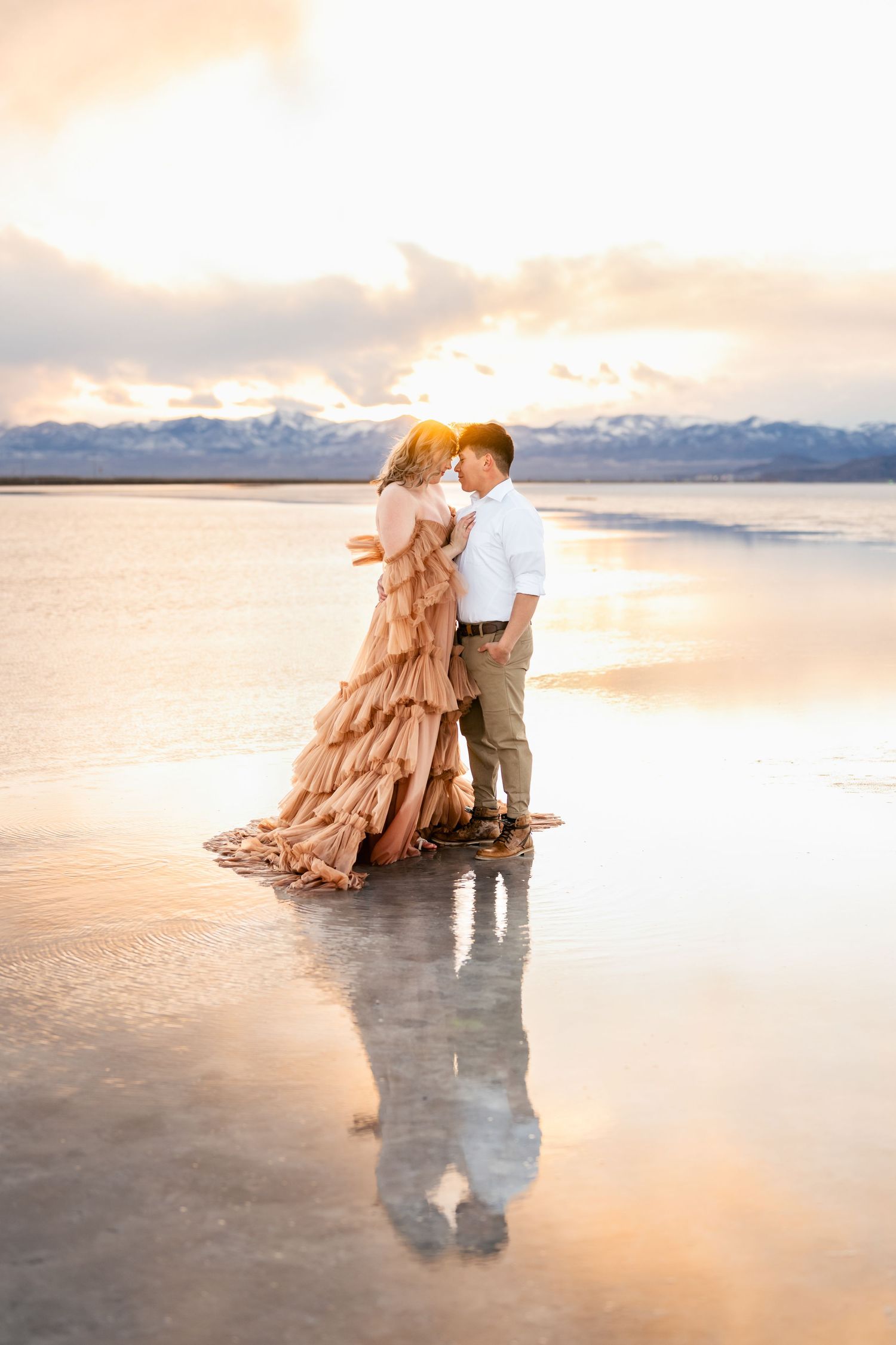 A couple stands forhead to forhead against the surreal backdrop of the flooded Salt Flats during golden hour.