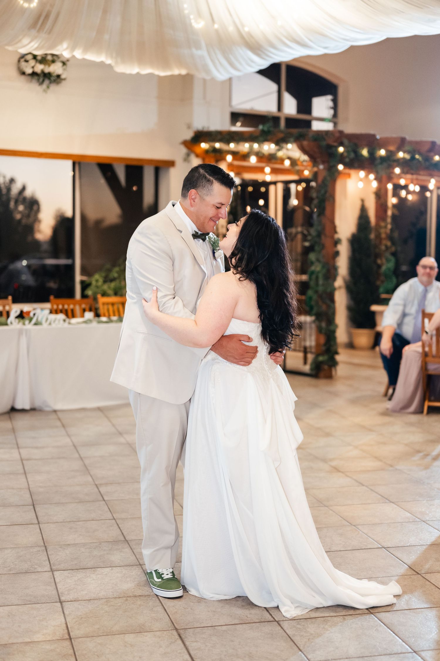 A couple shares an intimate dance in a wedding reception venue decorated with string lights and white drapes.