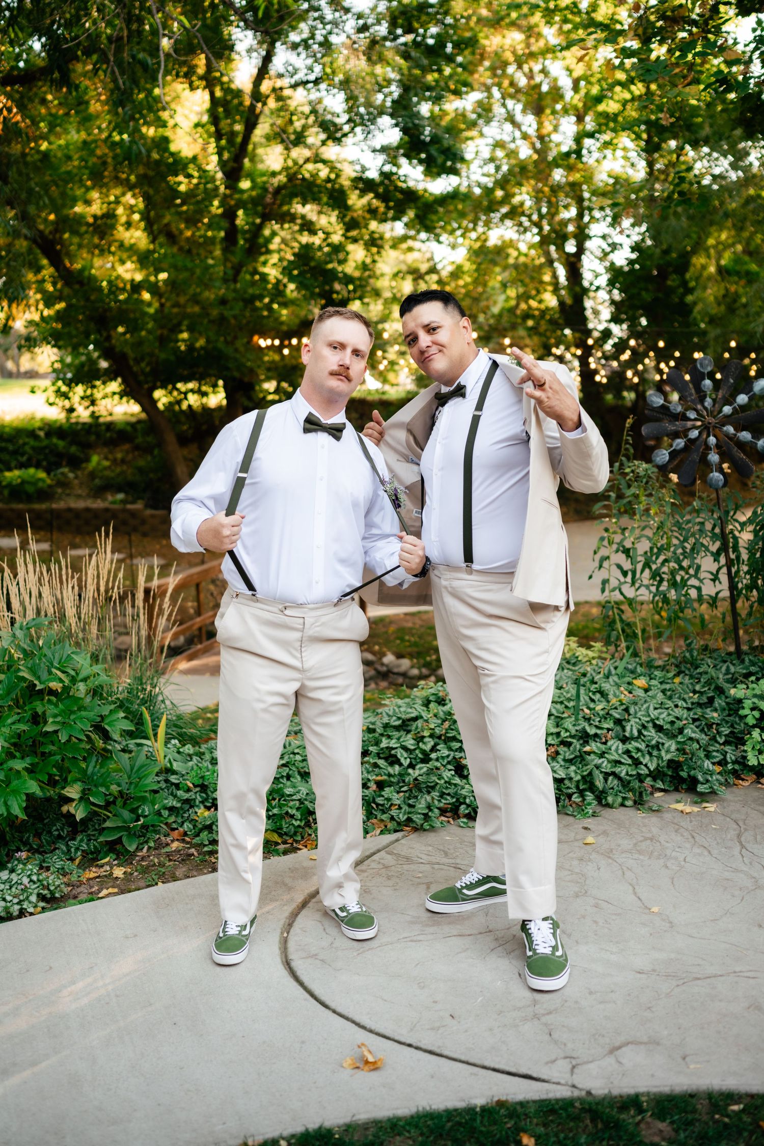 Two people in matching light-colored pants, suspenders and bow ties pose playfully on a garden path surrounded by greenery.
