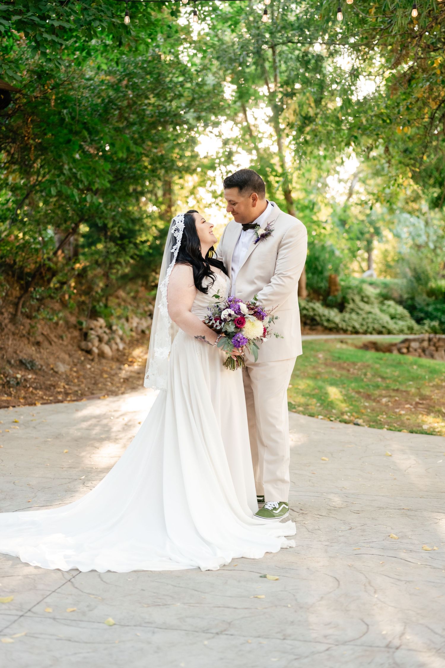 A couple in wedding attire share an intimate moment on a garden path, with the bride holding a purple and pink bouquet.