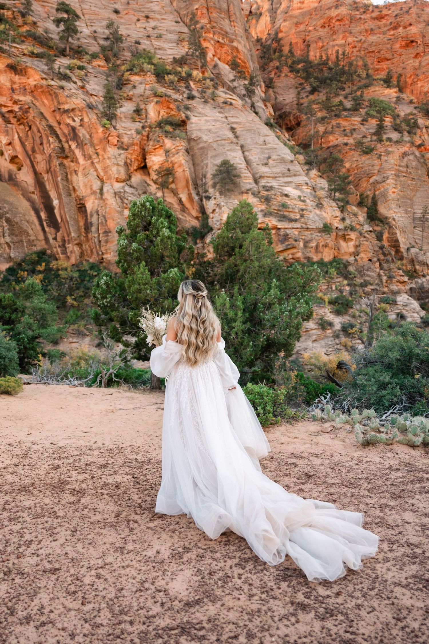 A flowing white wedding dress trails on dirt path surrounded by towering red rock formations.