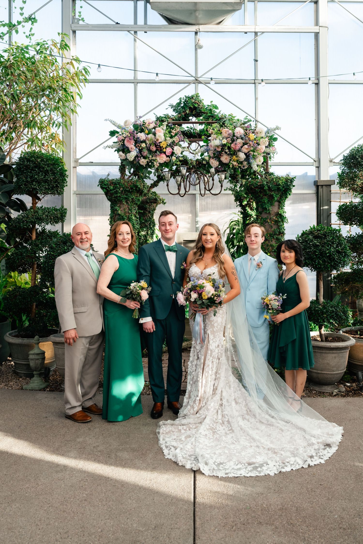 Wedding party poses together under a floral arch in a greenhouse with elegant dresses and flowers.
