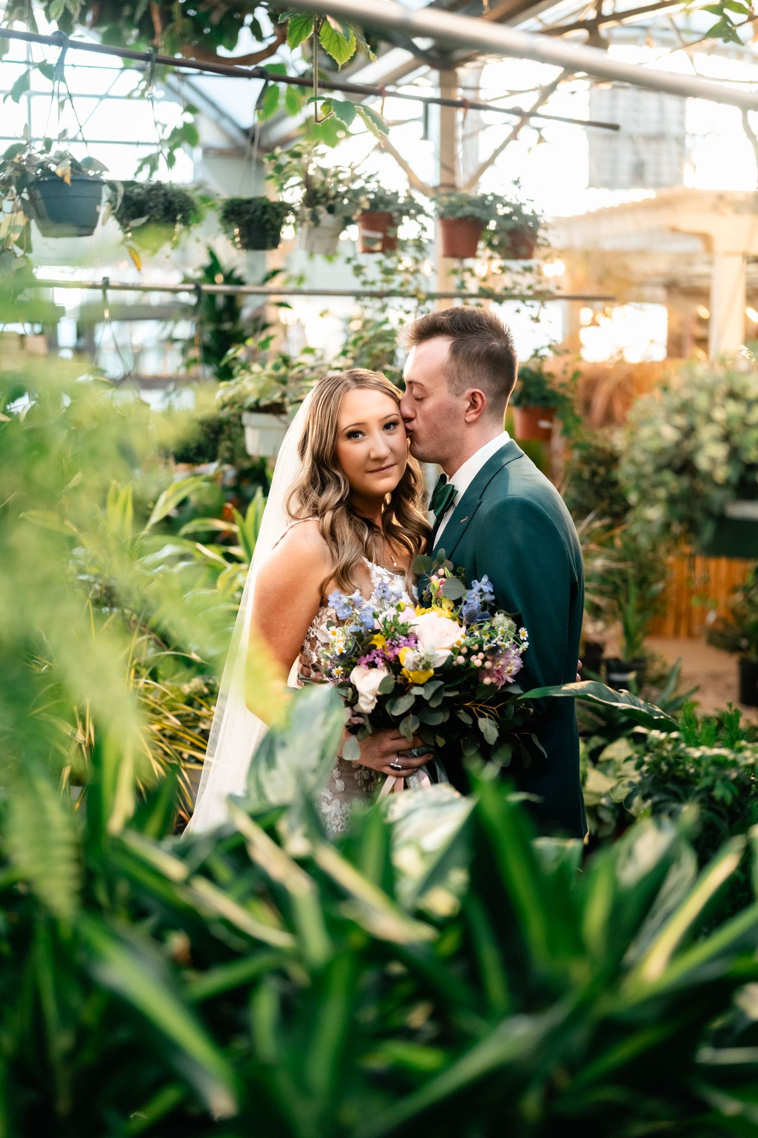 Intimate wedding portrait among lush greenhouse plants with natural sunlight filtering through.