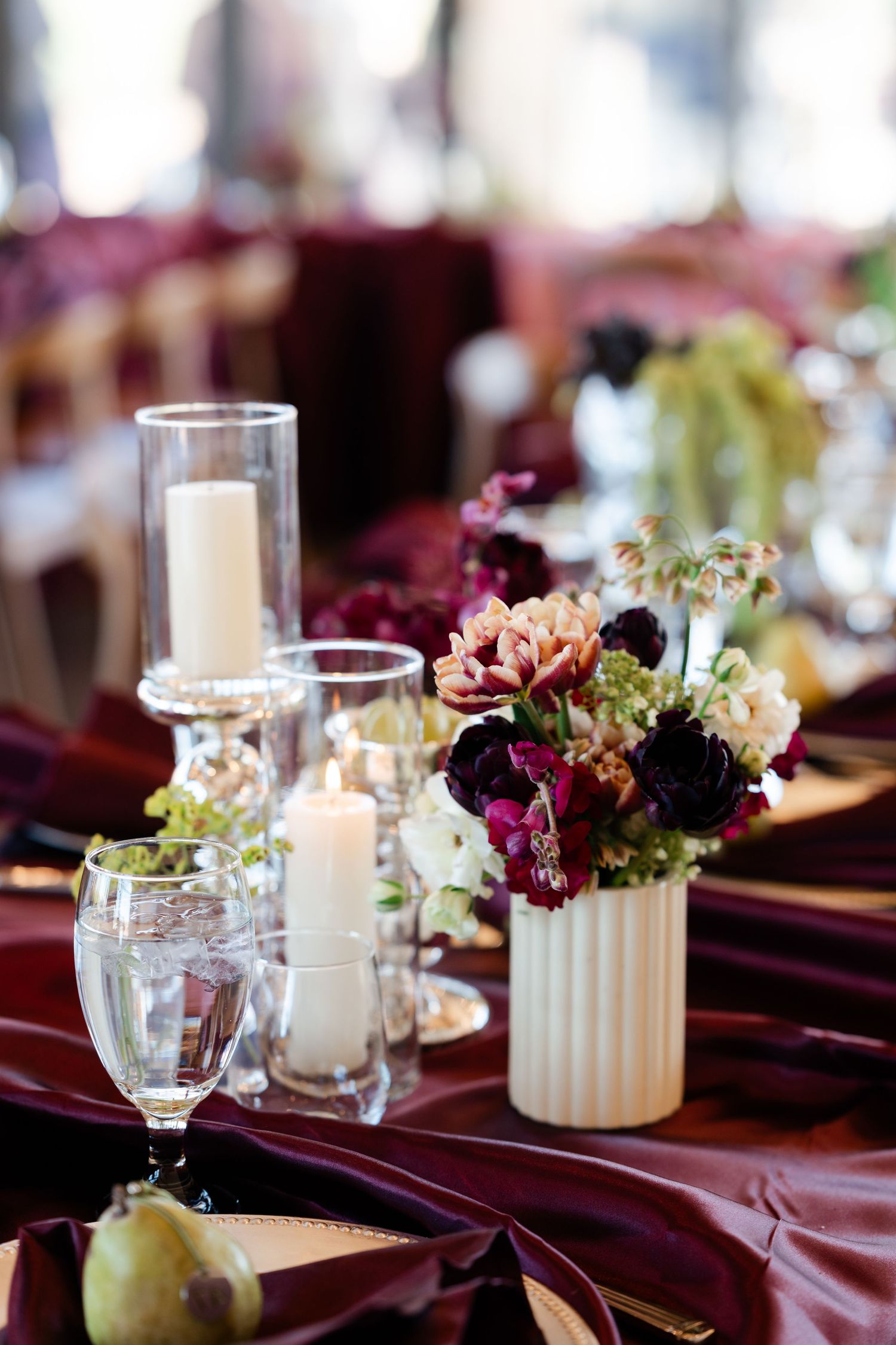 Elegant wedding reception table with burgundy linens, white pillar candles, and floral centerpiece featuring deep red and cream blooms.