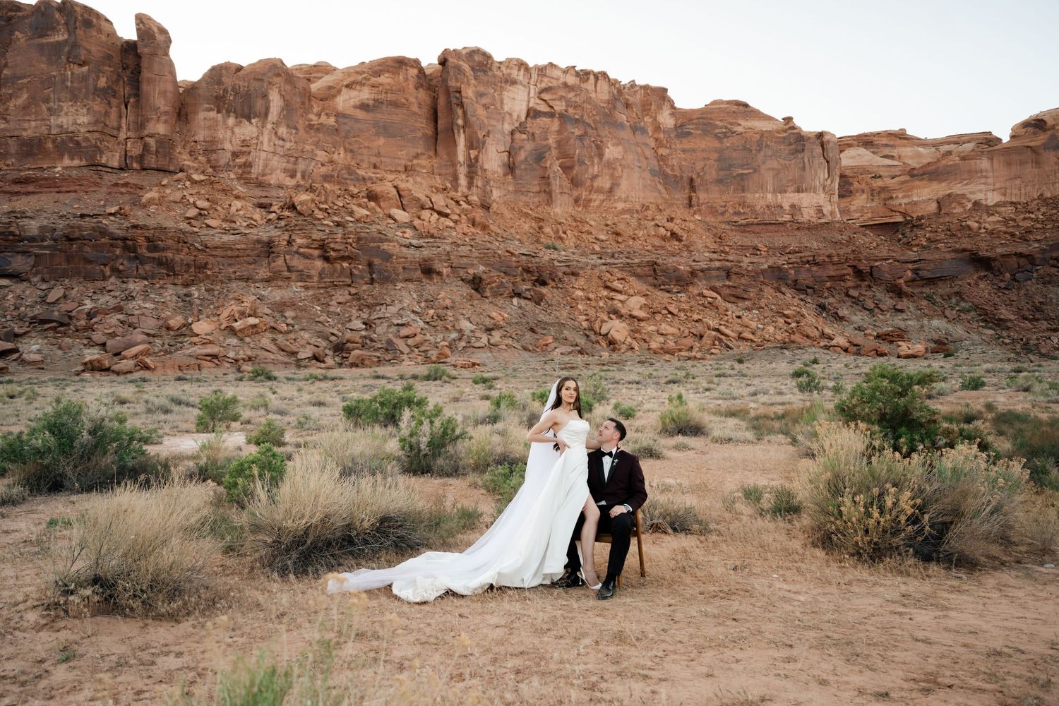 Bride in white gown and groom in tuxedo pose before red rock canyon formations in Moab.