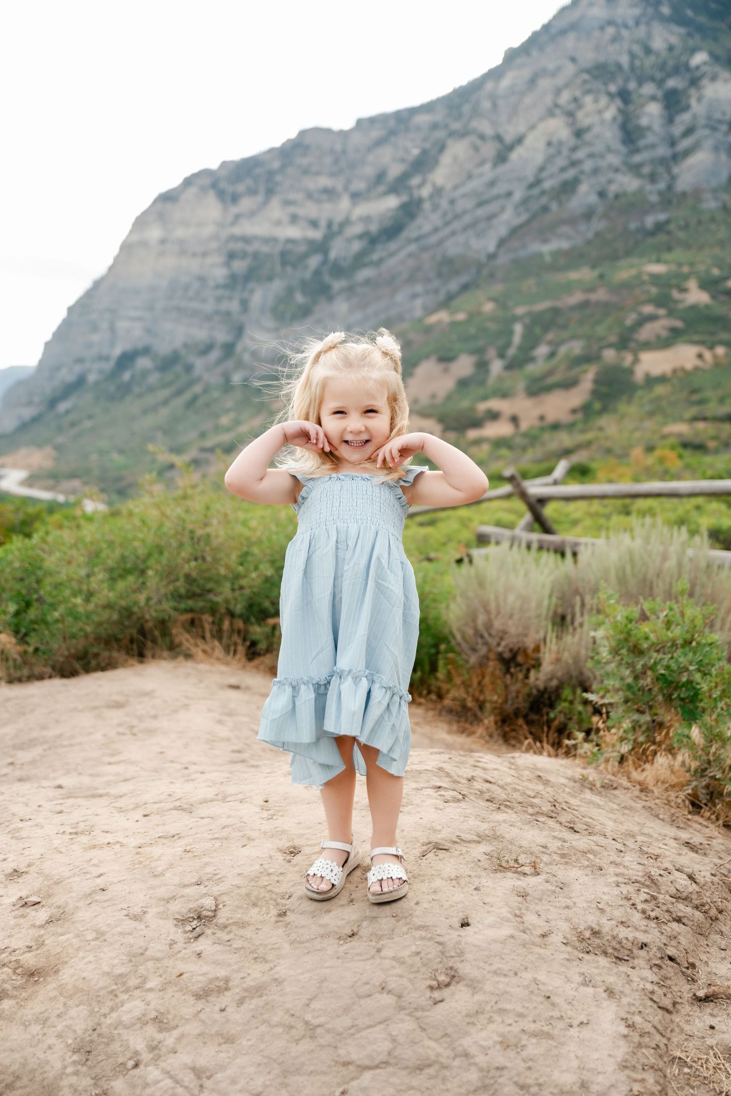 A child in a light blue dress plays joyfully on a rocky outcrop with rugged mountain scenery in the background.