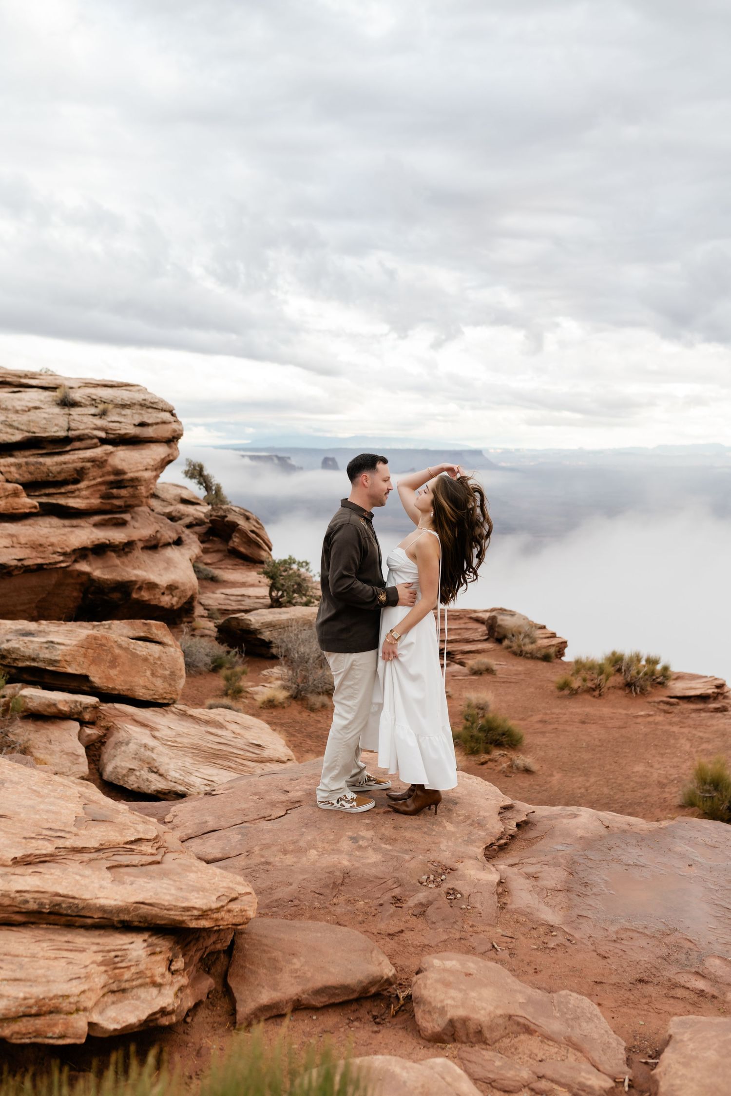 A couple stands together on red rock formations in a desert landscape under cloudy skies.