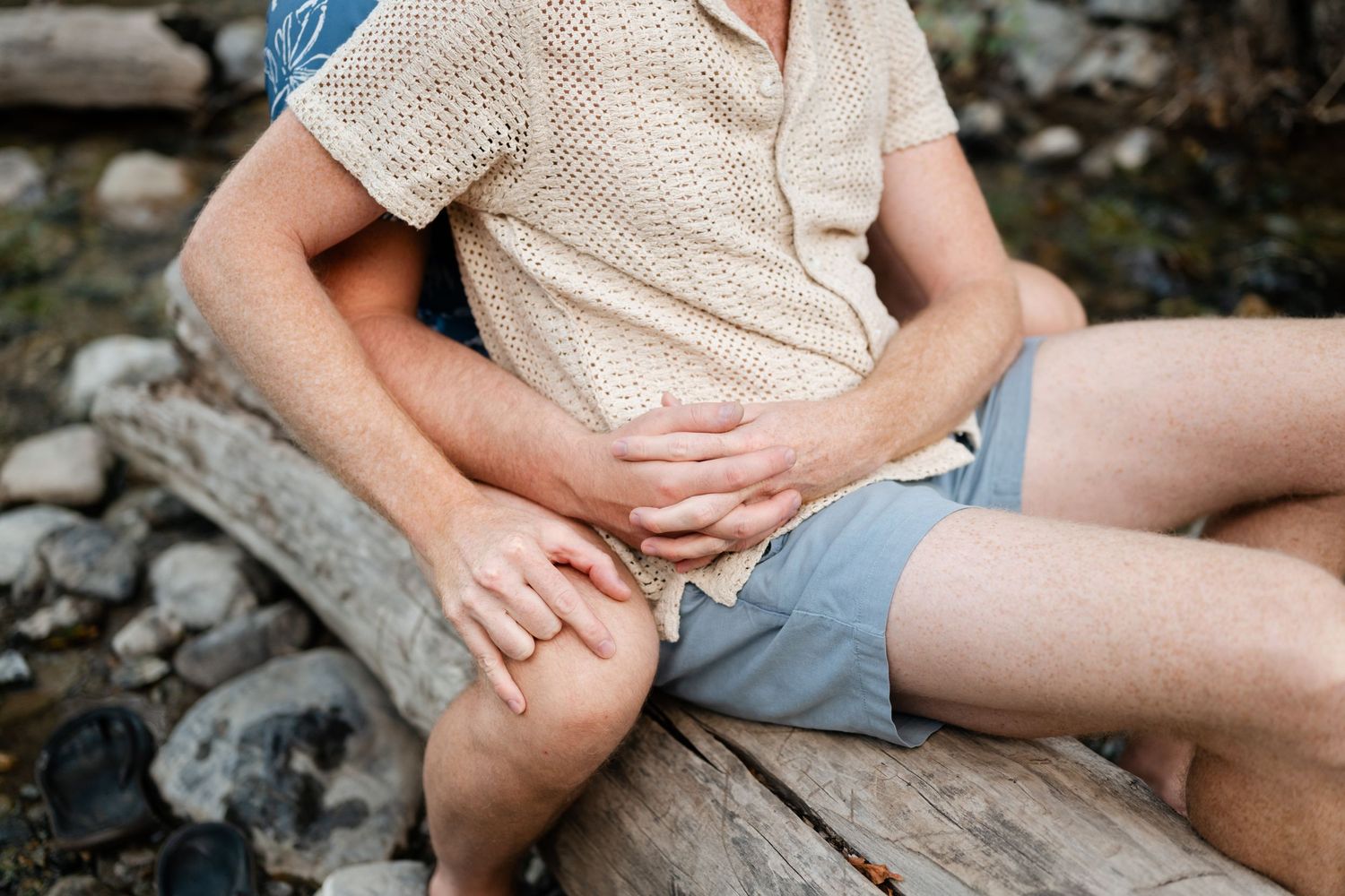 LGBTQ couple sitting together on a log outdoors, embracing near a rocky riverbed in casual summer clothing.