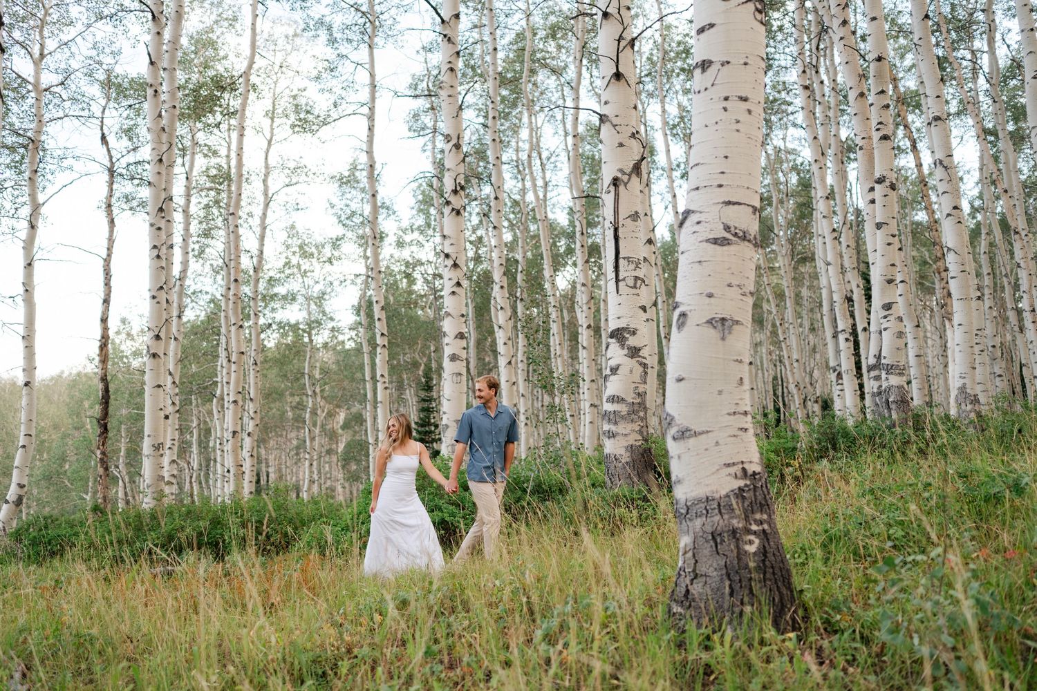 Bride and groom holding hands walking through a sunlit aspen forest grove during their outdoor Park City engagement.