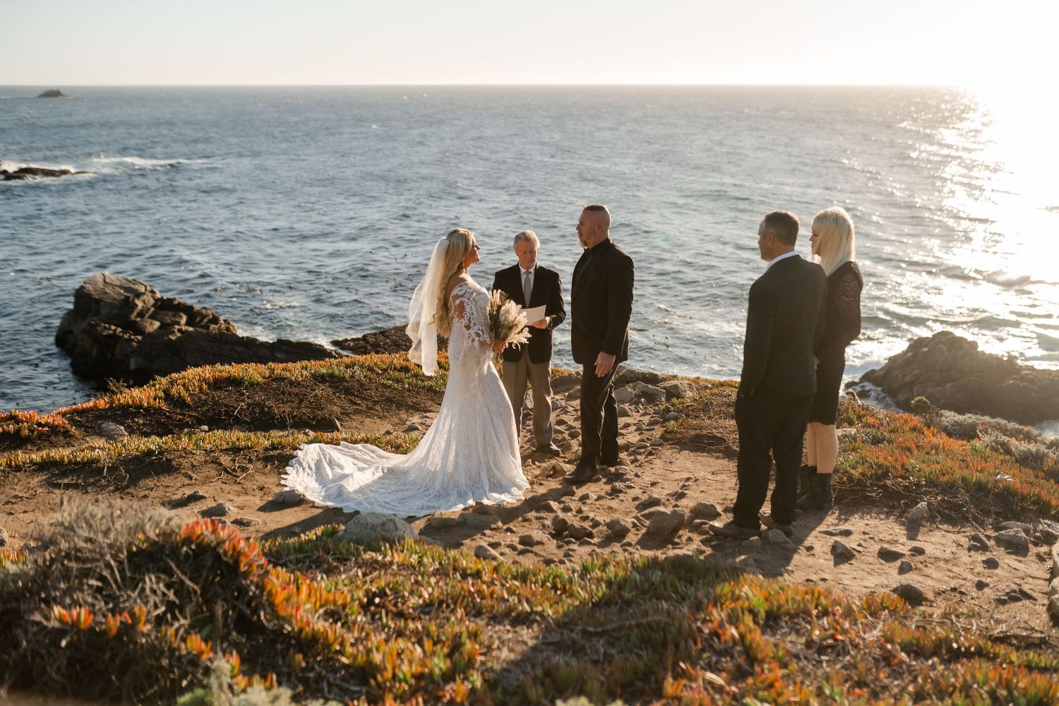 Seaside wedding celebration on dramatic cliffside overlooking Pacific Ocean during sunset.
