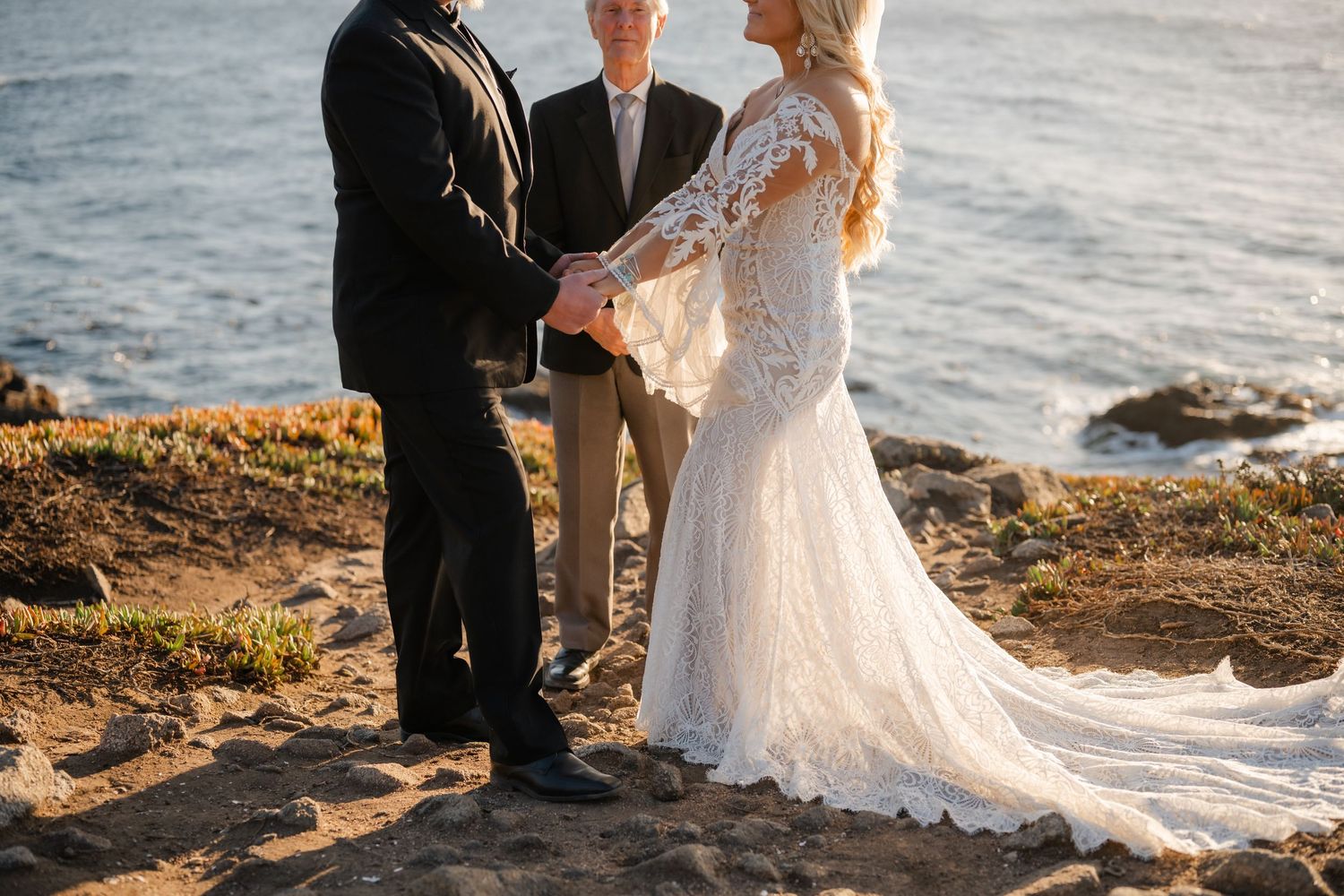 A couple in wedding attire stands on a rocky coastal cliff during golden hour as waves crash behind them.