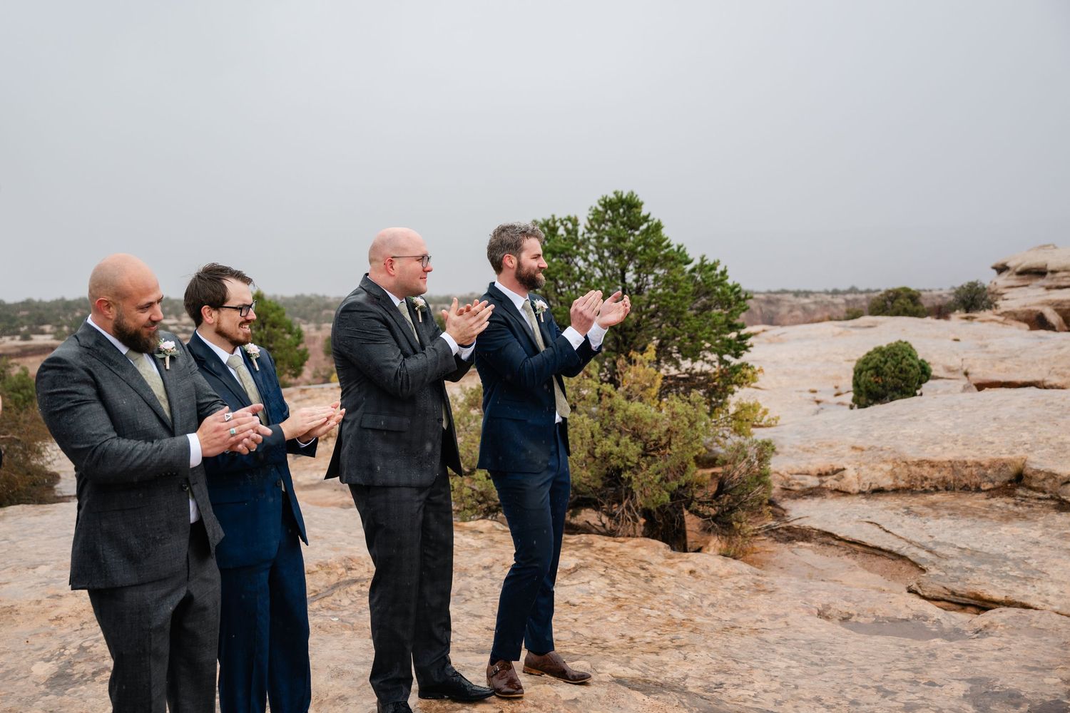 A group of people in formal attire stand on a rocky desert landscape during an overcast day.