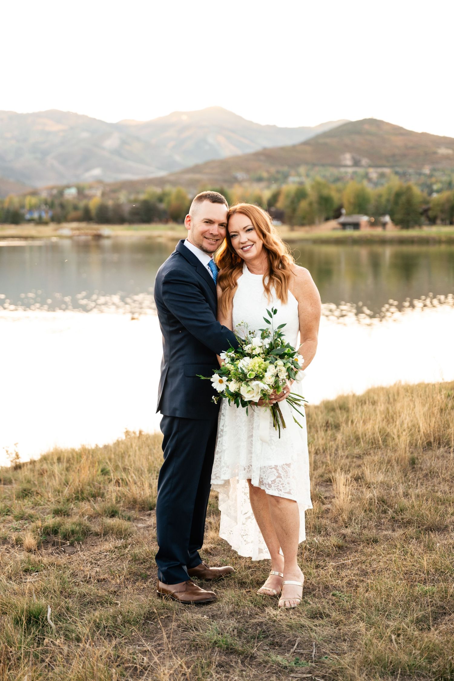 A couple poses together near a lake with mountains and golden light in the background.
