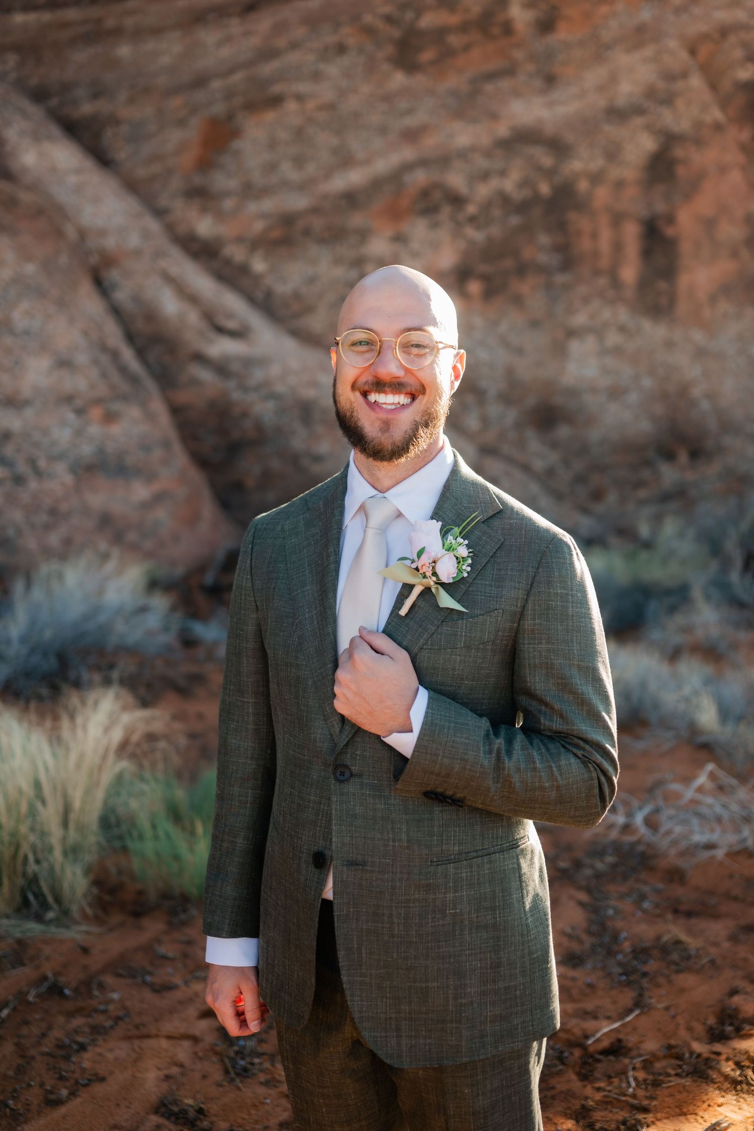 A groom in a gray suit with boutonniere poses in front of desert red rocks.