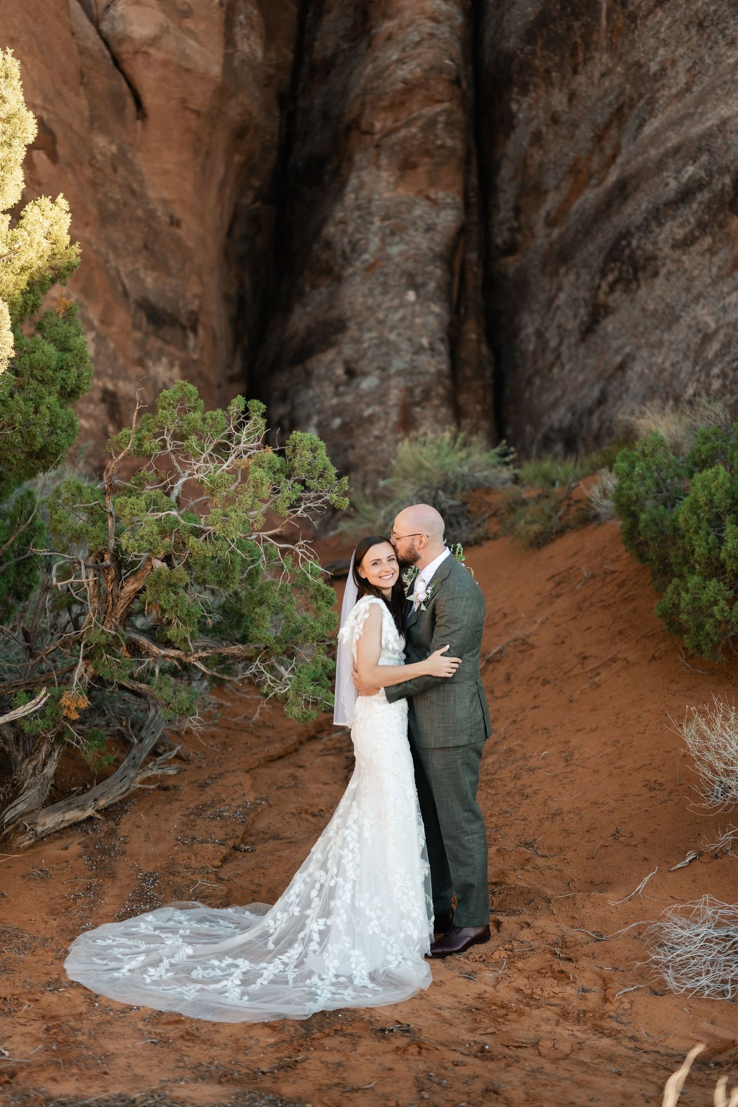 A couple in wedding attire embraces on a desert trail surrounded by red rock cliffs.