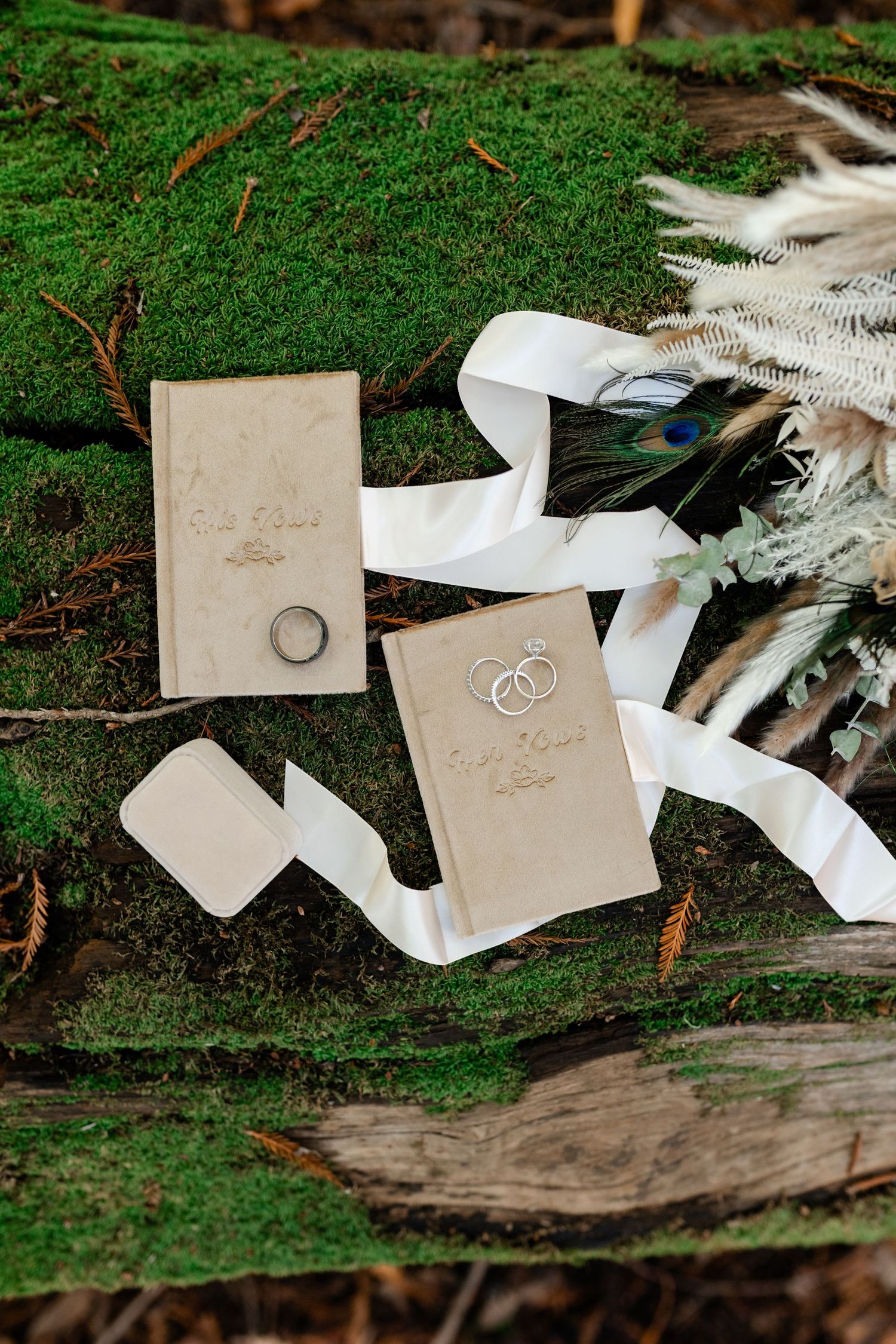 Wedding vows and rings displayed on moss with white feathers in a natural outdoor setting.