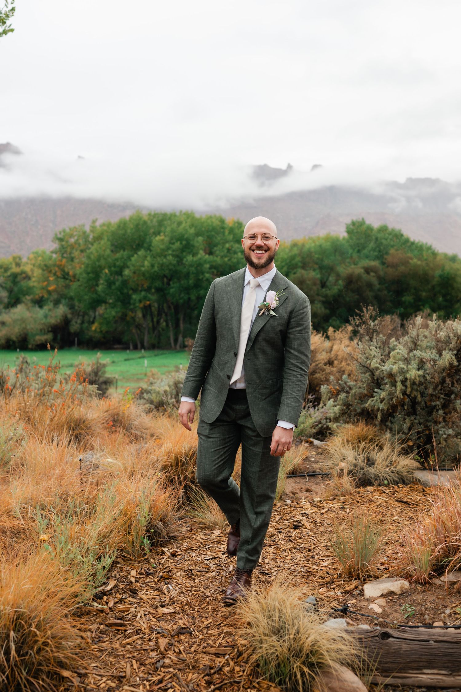 A person in a dark suit poses in a desert landscape with mountains and tall grasses.