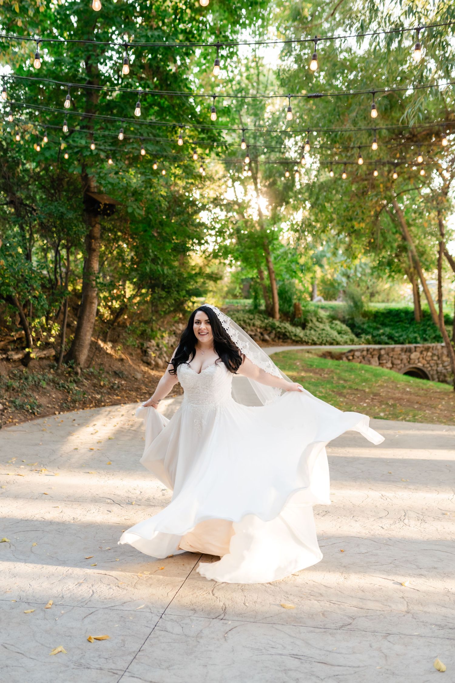 A bride in a flowing white dress twirls on a sunlit garden path under string lights and trees.