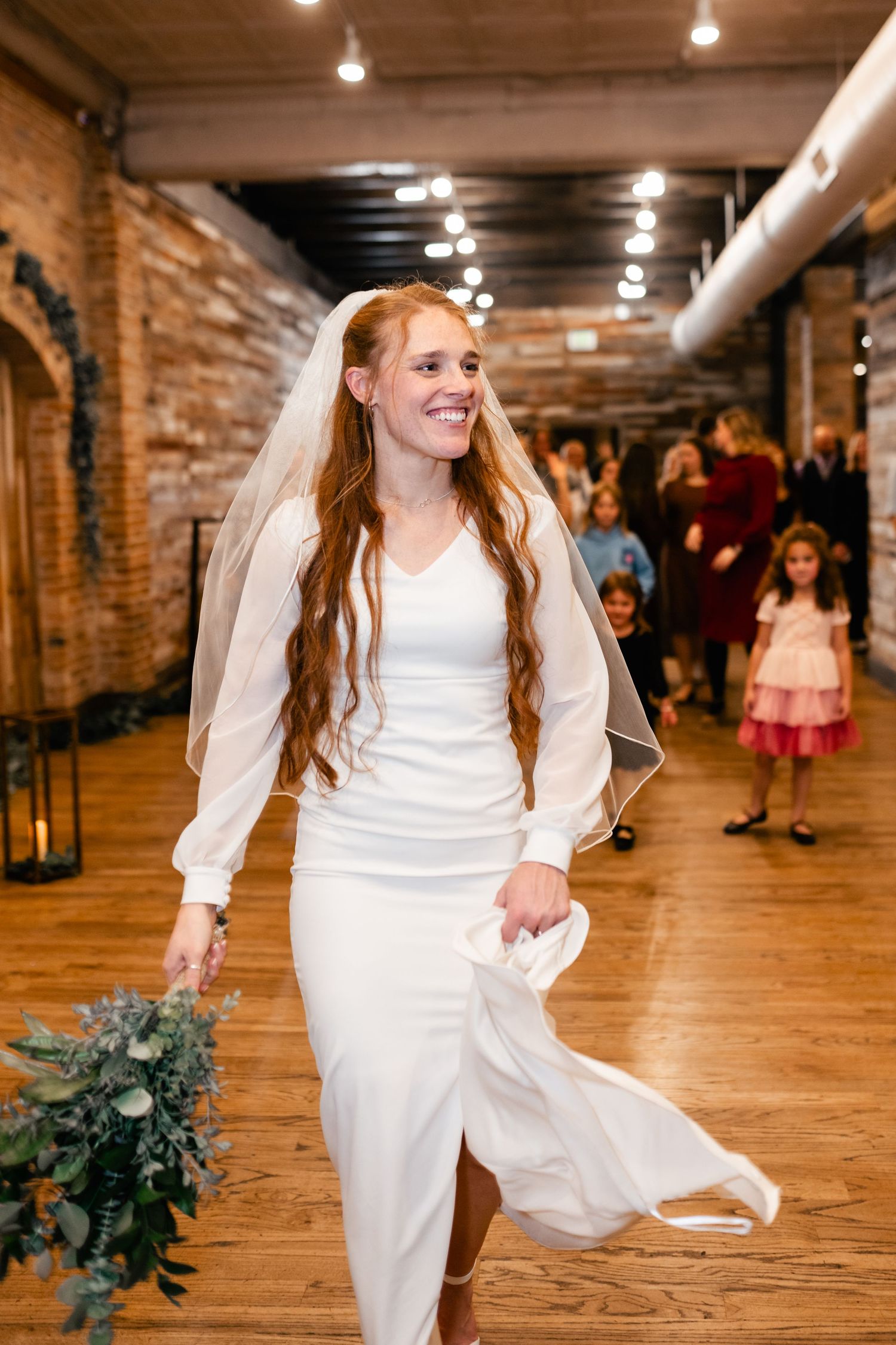 A bride in a long-sleeved white wedding dress dances on a hardwood floor in a brick-walled venue with string lights overhead.