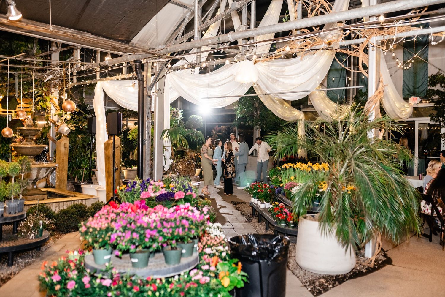A dreamy garden nursery interior decorated with white fabric draping, tropical plants and vibrant pink flowers for sale.