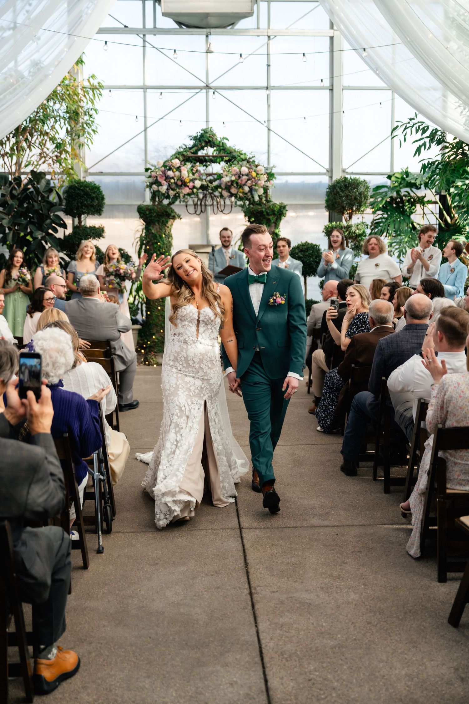Wedding ceremony recessional in a greenhouse venue with floral arch and seated guests.