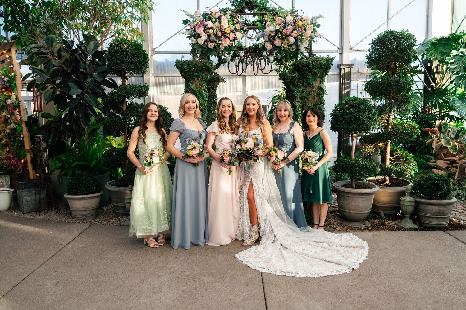 Bridal party in mix of sage green, blue and emerald dresses pose with bride in greenhouse setting with potted plants.