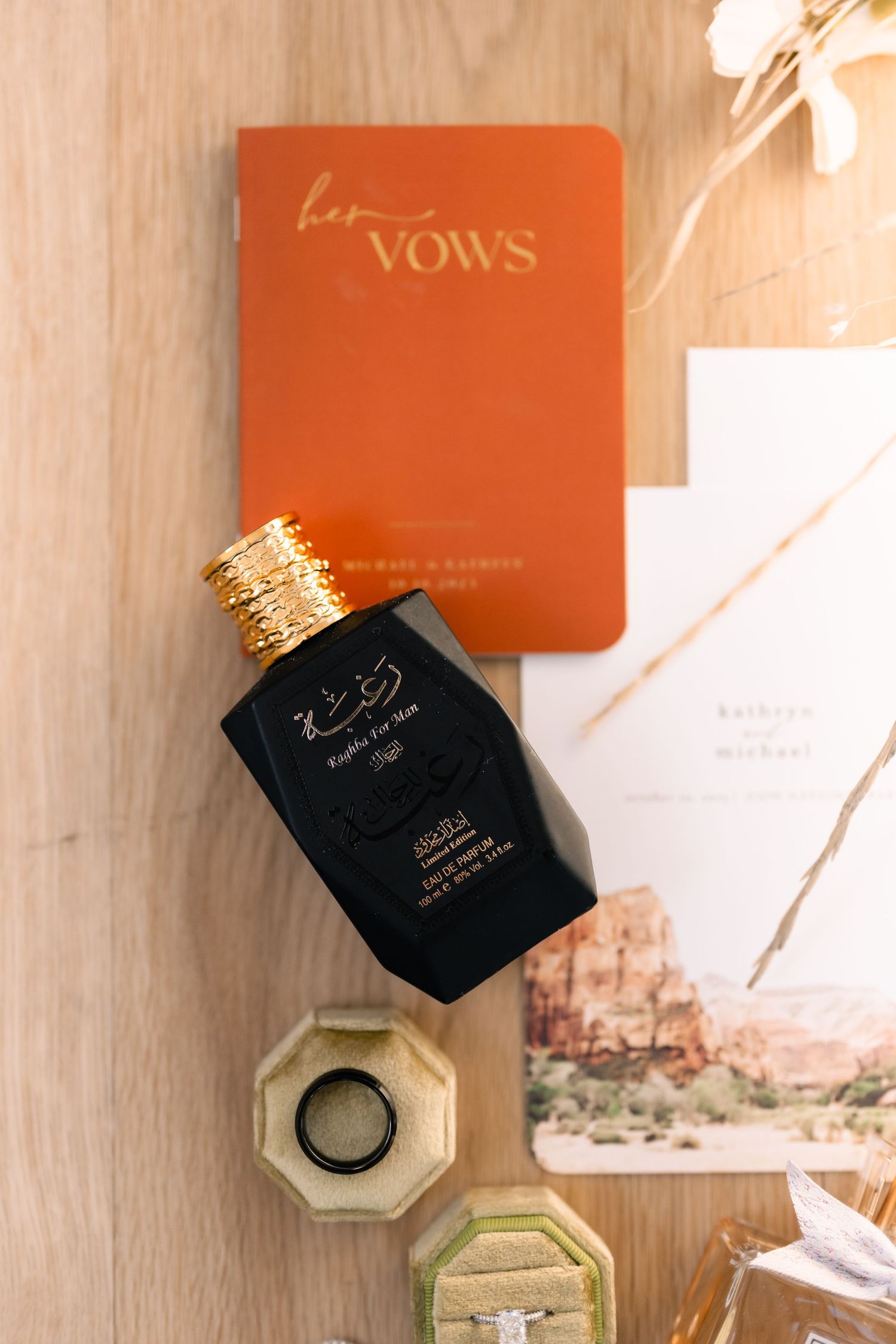 Wedding vow book and perfume bottle arranged with desert-themed invitation on wooden surface.