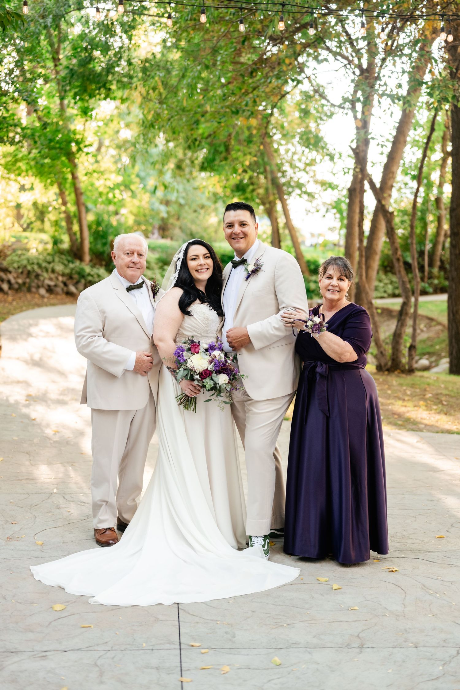 A wedding party poses outdoors wearing light colored and purple formal attire with the bride holding a vibrant bouquet.