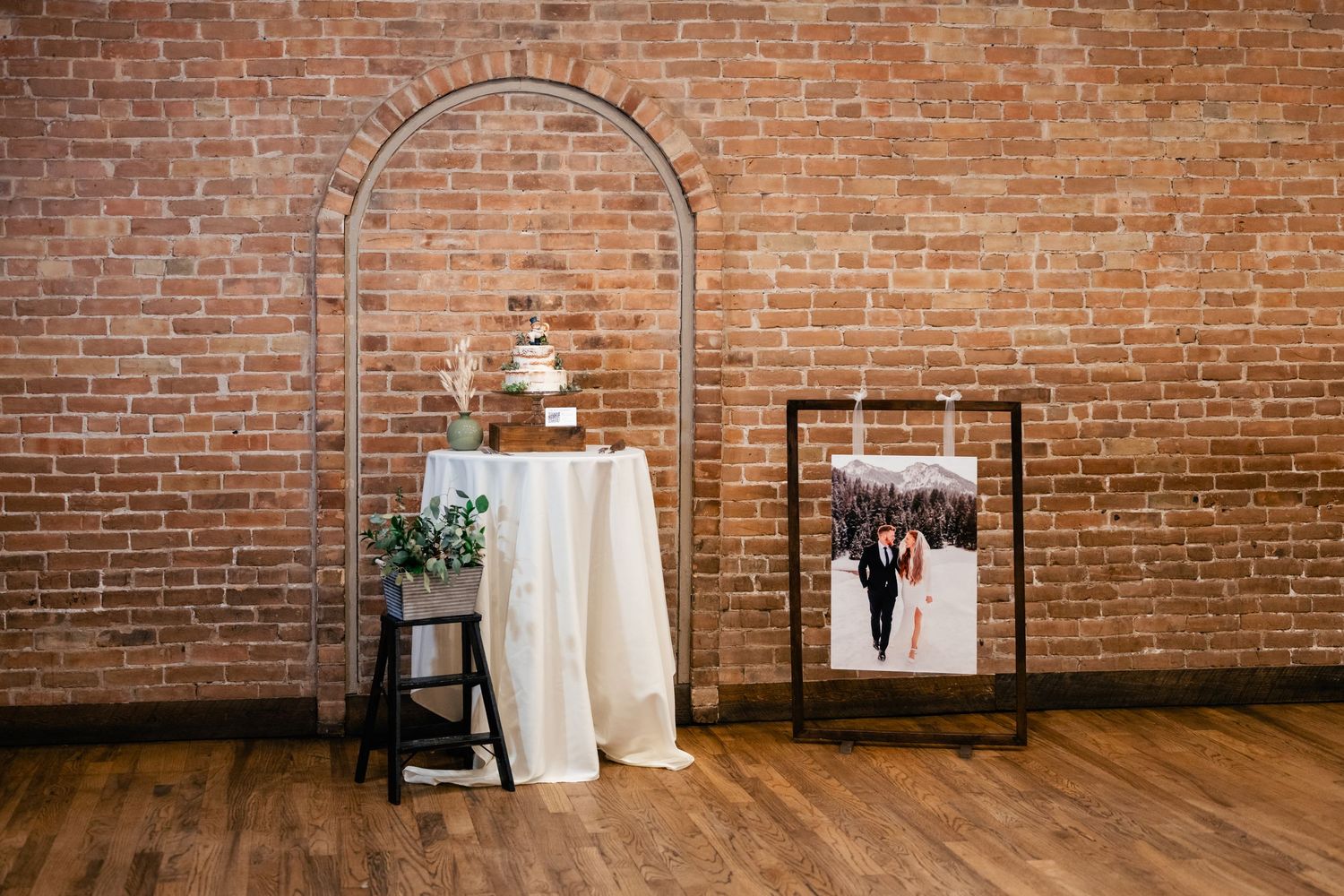 Wedding display with tiered white cake and framed photo against exposed brick wall and metal arch in rustic venue.
