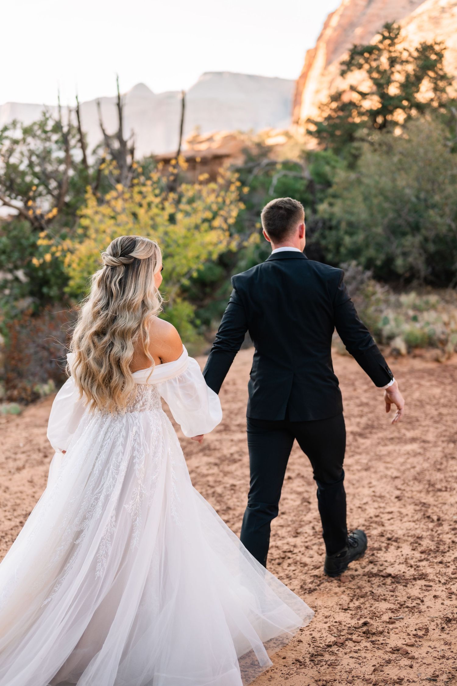 Wedding couple strolling through desert terrain with flowing white dress and dramatic cliffs.