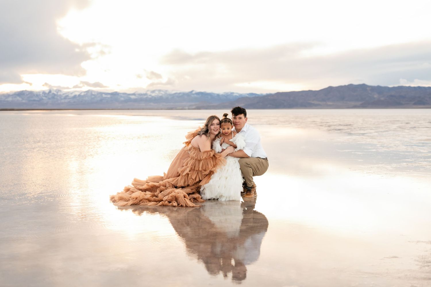 Parents embrace their child affectionately amidst the vast expanse of the flooded salt flats during golden hour.