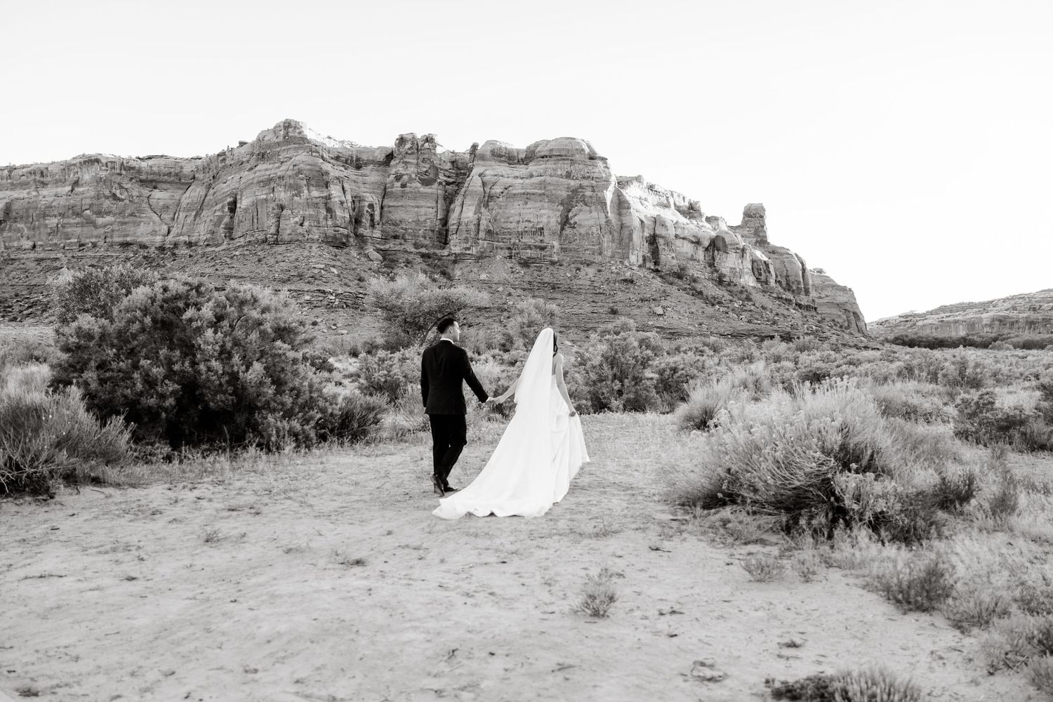 Black and white photo of bride and groom walking in desert landscape with dramatic mesa rock formations behind them.