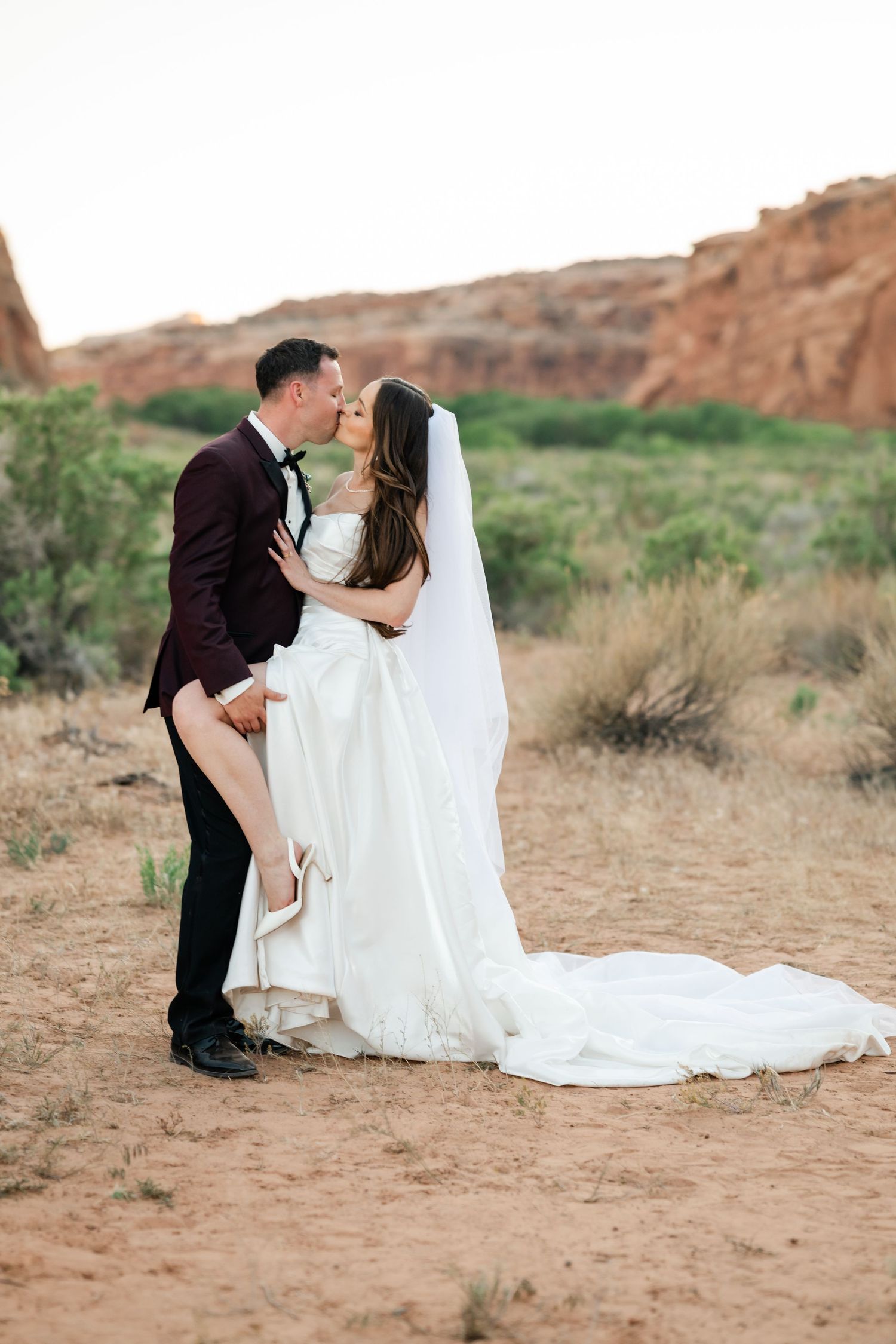 Bride and groom share a romantic kiss in a desert landscape with red rock formations, bride in white gown with veil.