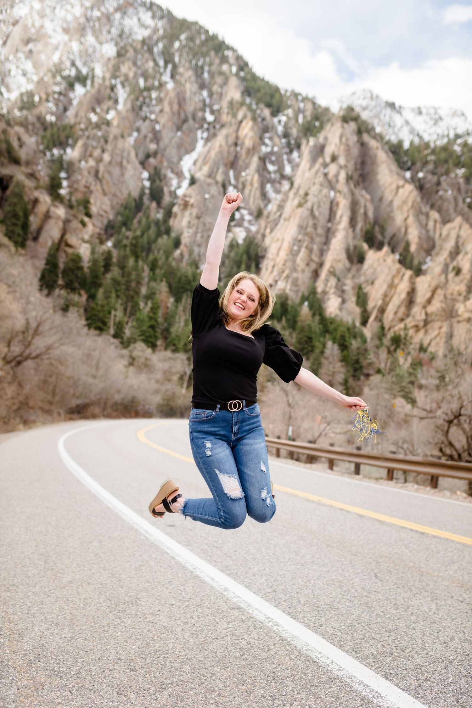 A person in jeans and black top jumps and poses playfully on a mountain road with dramatic rocky cliffs in the background.
