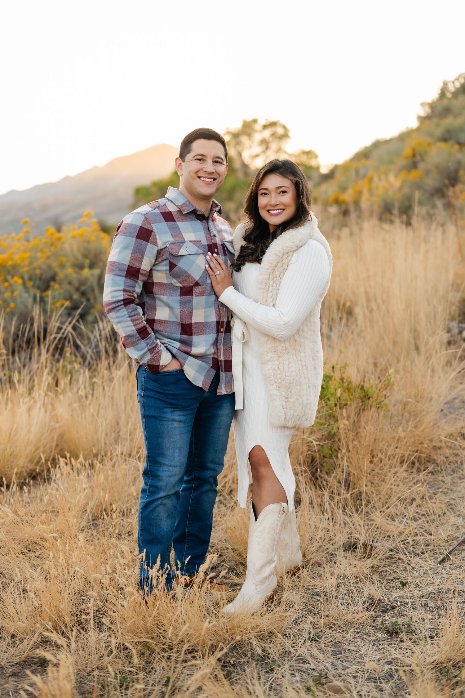 A couple poses together on a hillside during golden hour wearing casual fall outfits and white boots in autumn weather.