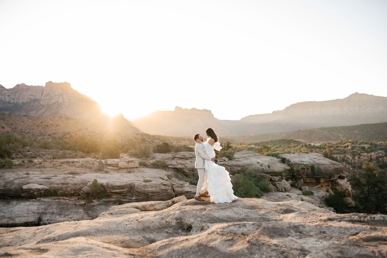 Zion Adventure Session. Wedding photos captured on rocky desert overlook during golden hour with mountains in distance.