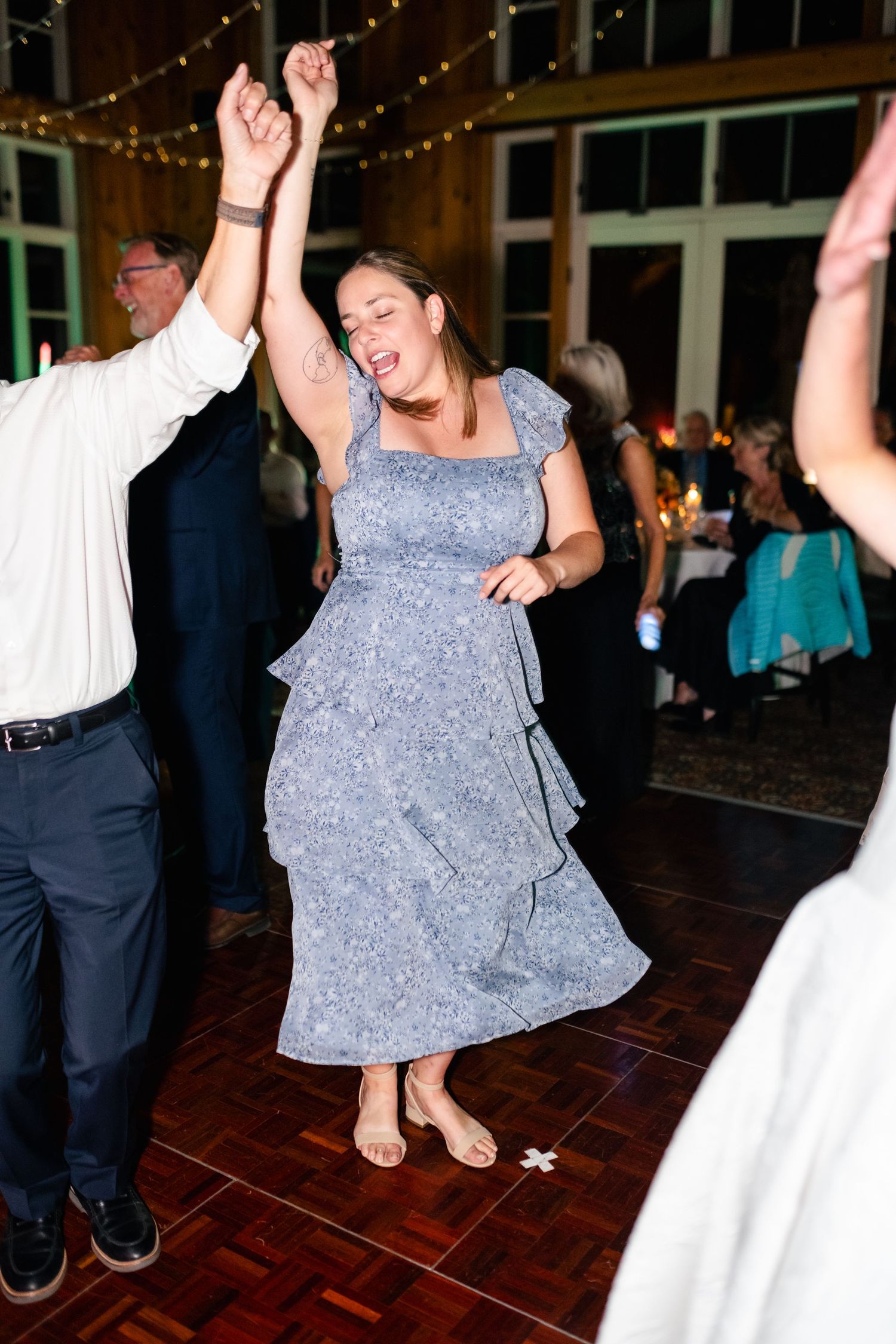 Wedding guests dancing energetically on dark wooden dance floor under string lights.