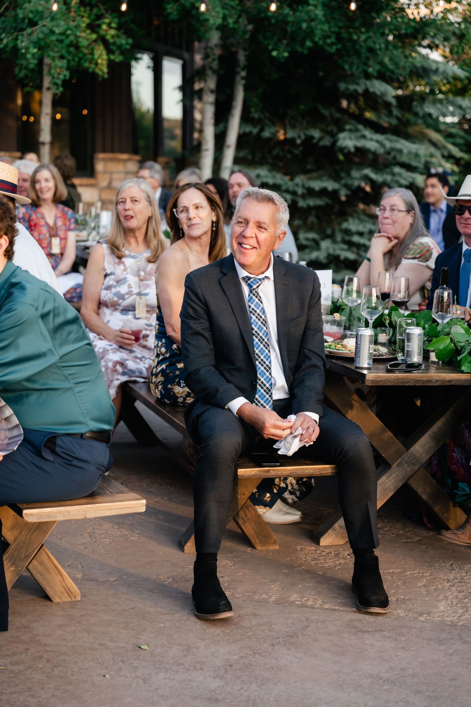 Guests enjoy an outdoor wedding reception at rustic wooden tables under string lights and trees.