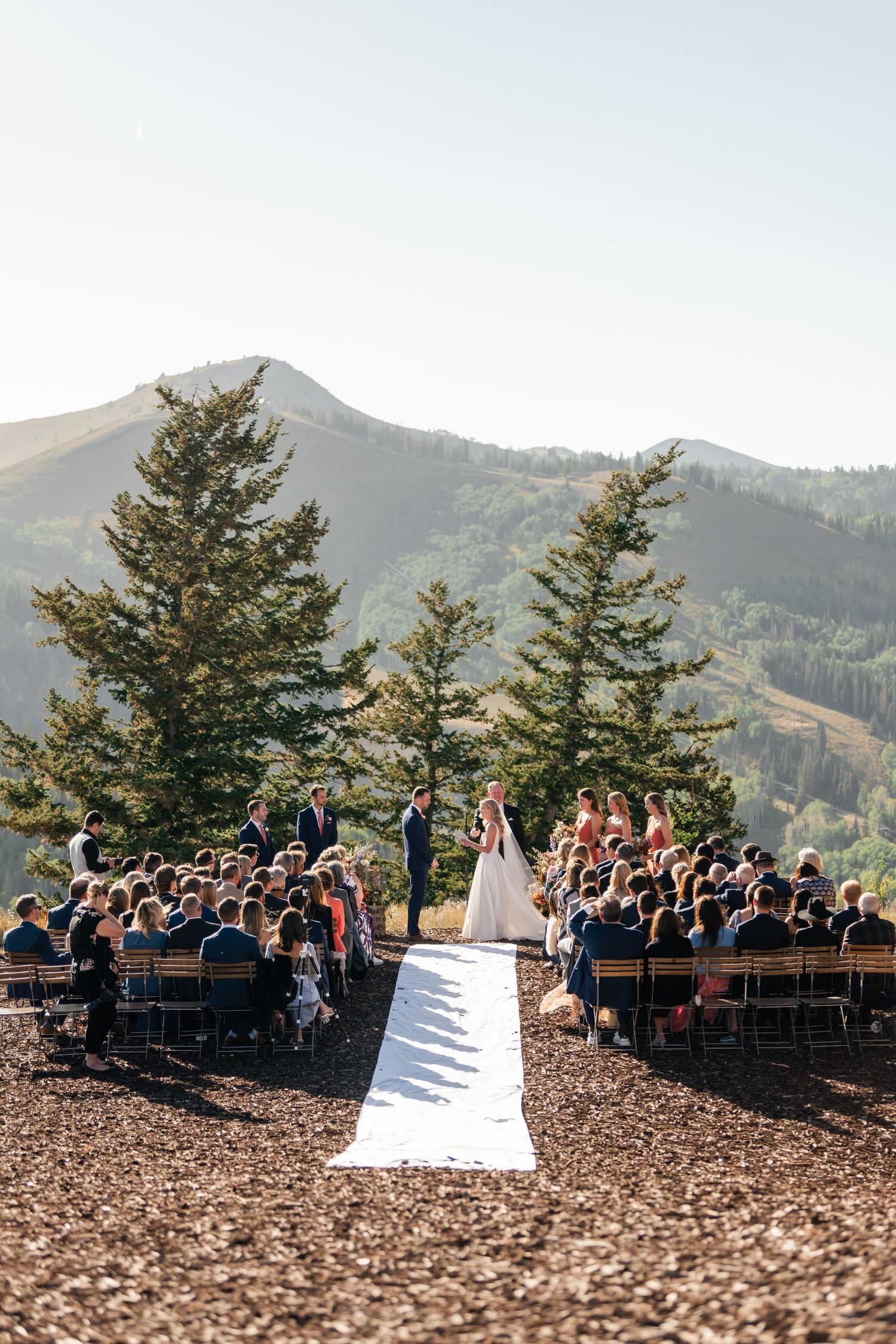 A scenic outdoor mountain wedding ceremony with guests surrounded by evergreen trees. Mountain top ceremony in Deer Valley, Utah.