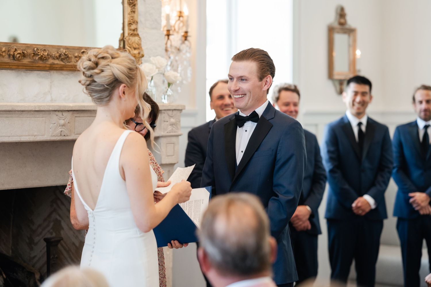 Elegant wedding ceremony takes place in a bright room with classic decor and ornate gold-framed mirror.