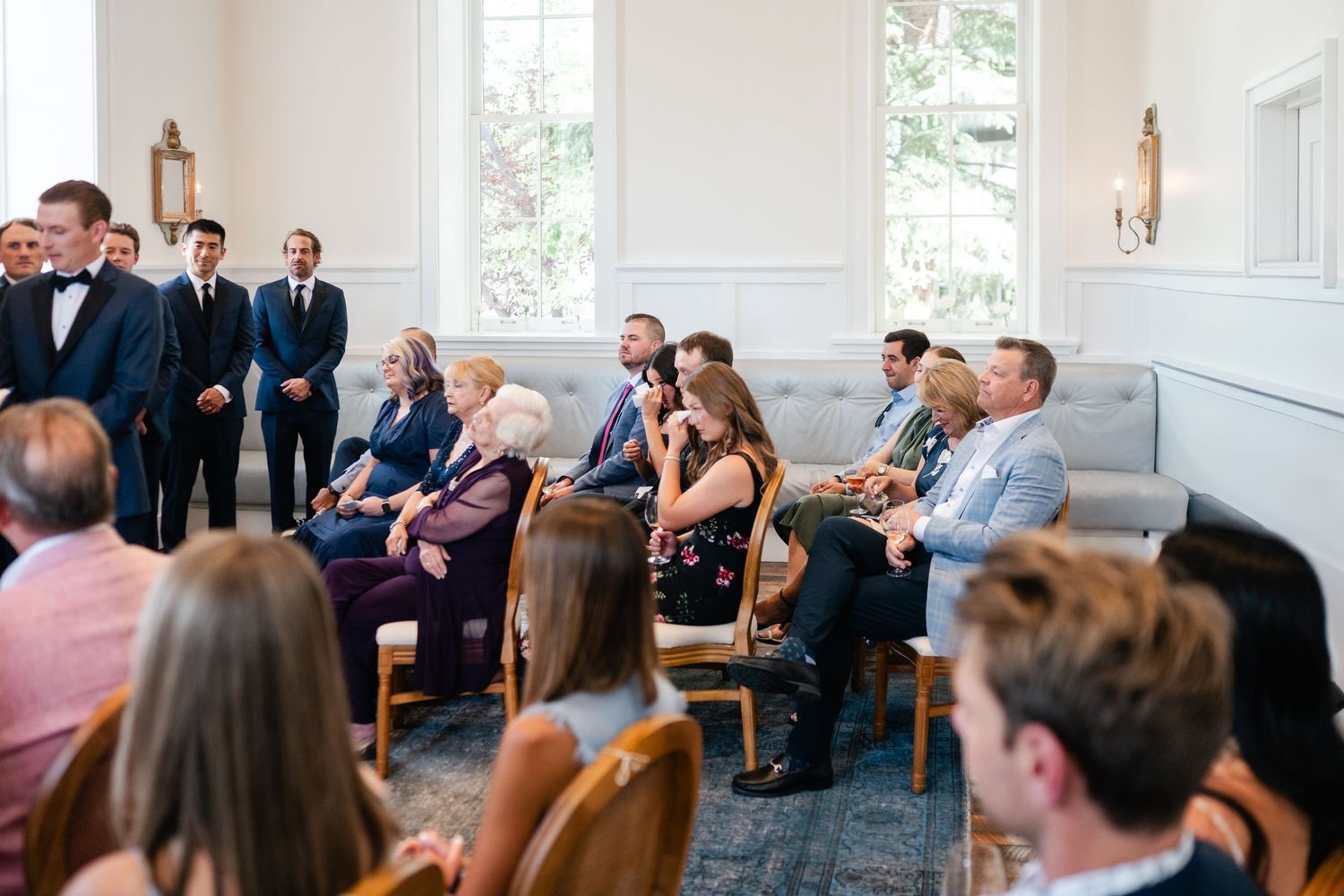 Wedding guests seated in white church pews watch attentively during an indoor ceremony.