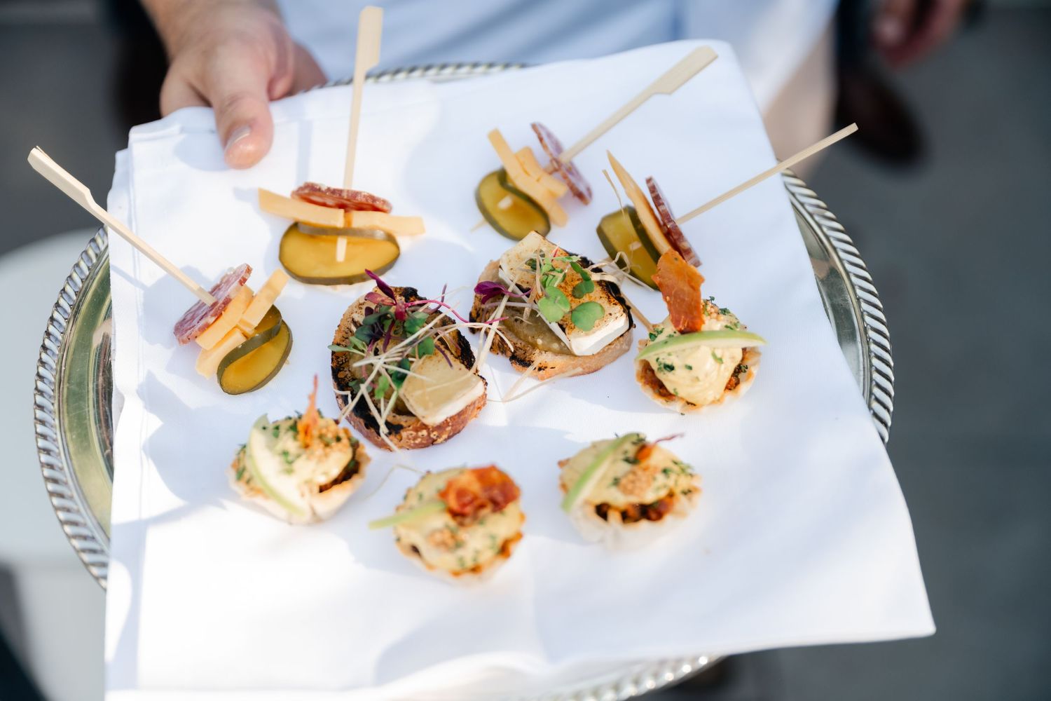 Hands reaching for gourmet mini sliders and appetizers served on a white platter at an elegant event.