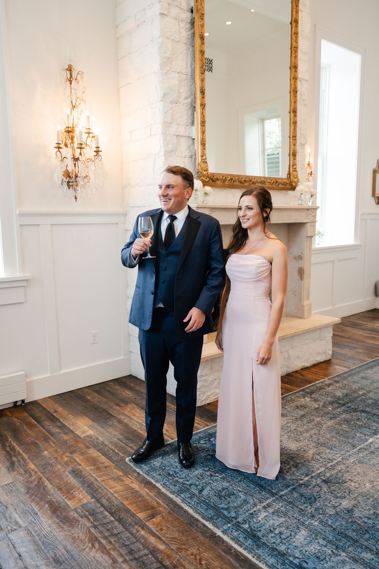 Elegant couple poses in formal attire against ornate wall with chandeliers in vintage-style indoor setting.