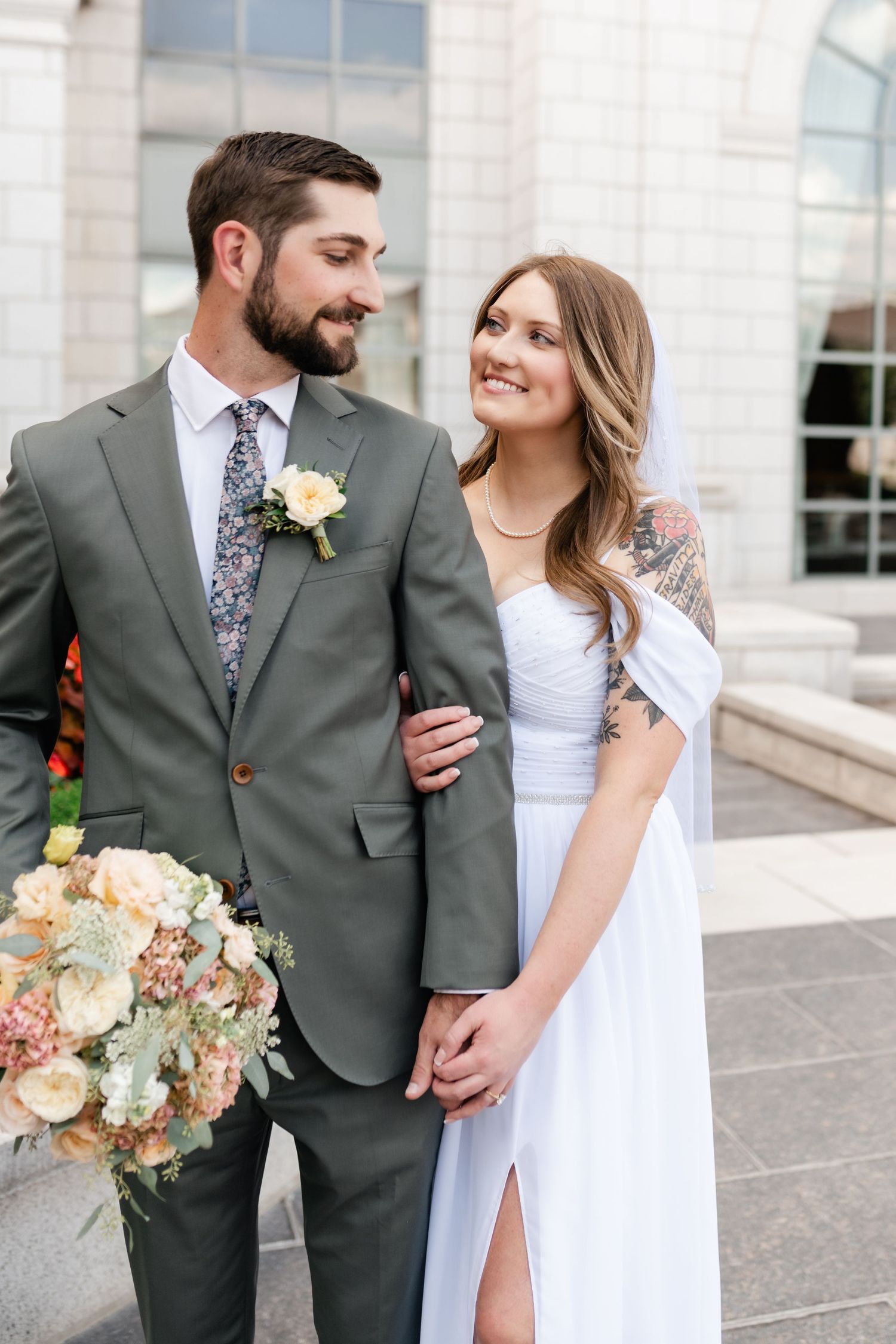 A couple in wedding attire stands together outside a building, with the bride in white and groom in gray suit.