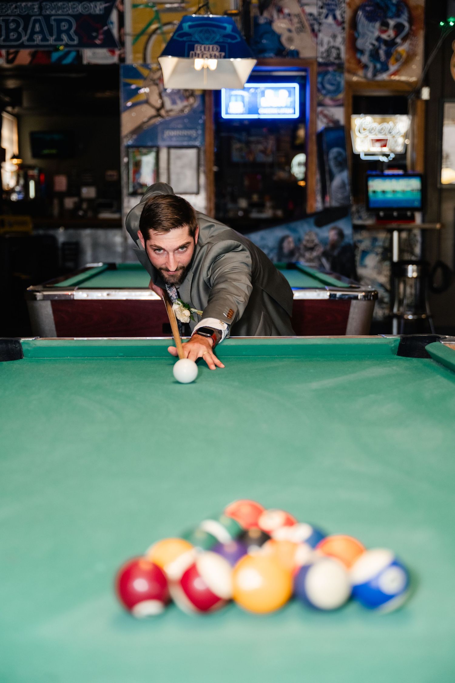 A person in a gray sweater lines up a shot while playing pool on a green felt table in a bar setting.