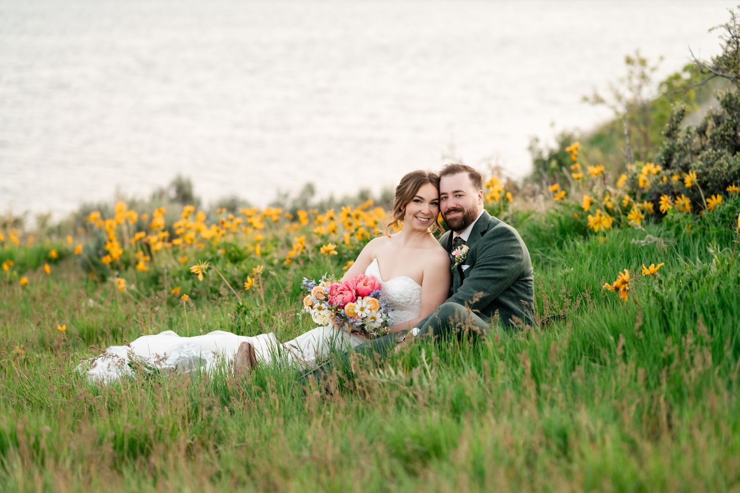 Couple embraces in field of yellow wildflowers with mountain lake backdrop.