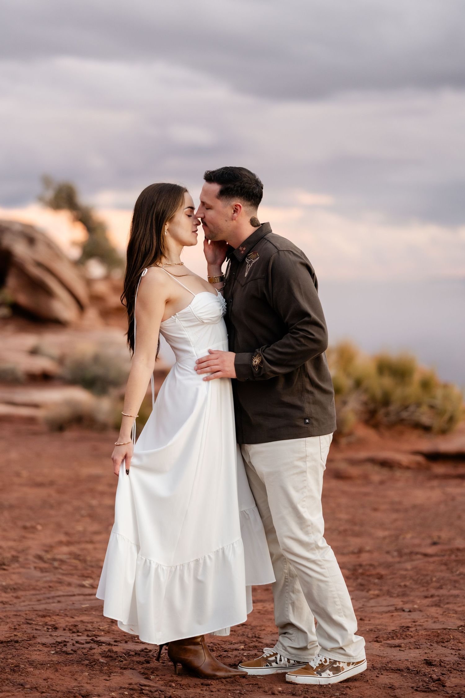 A romantic couple embraces on red rocks during sunset wearing white dress and brown jacket.